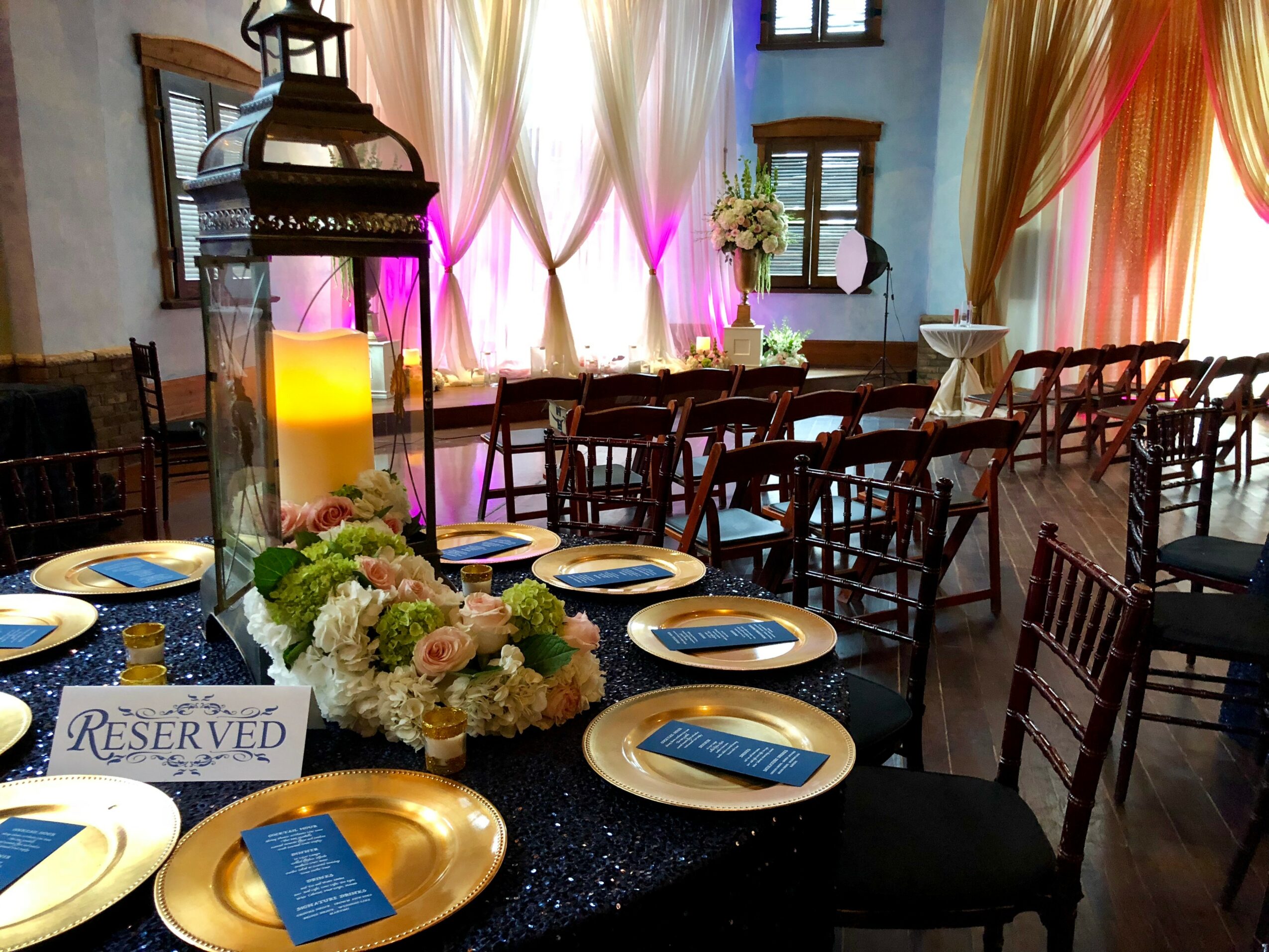 Elegant event setup with reserved sign, floral centerpiece, gold plates, and draped curtains in a decorated room.