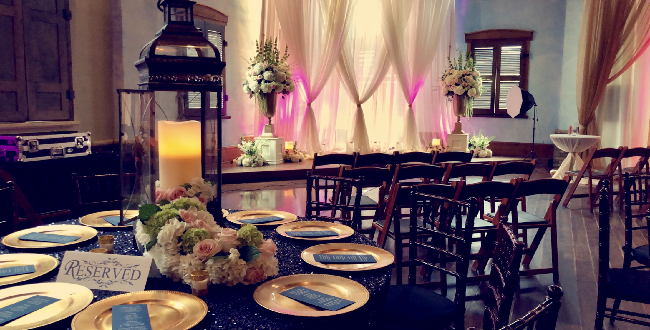 Elegant dining table set with gold-rimmed plates, floral centerpiece, and candles, in a decorated room with pink curtains and flowers.