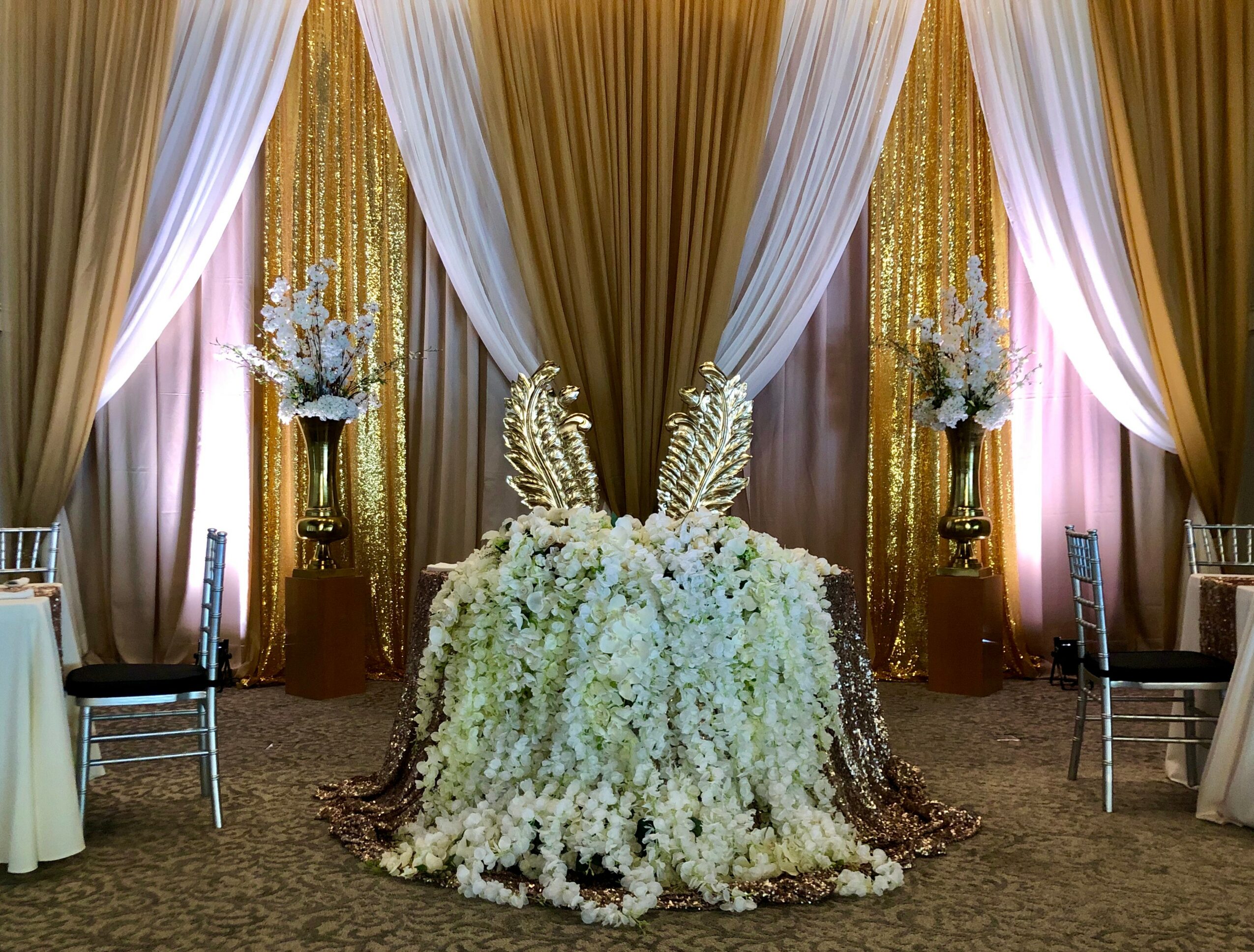 Elegant floral arrangement with white flowers and decorative leaves on a table, backdrop of gold and white drapes with lighting.