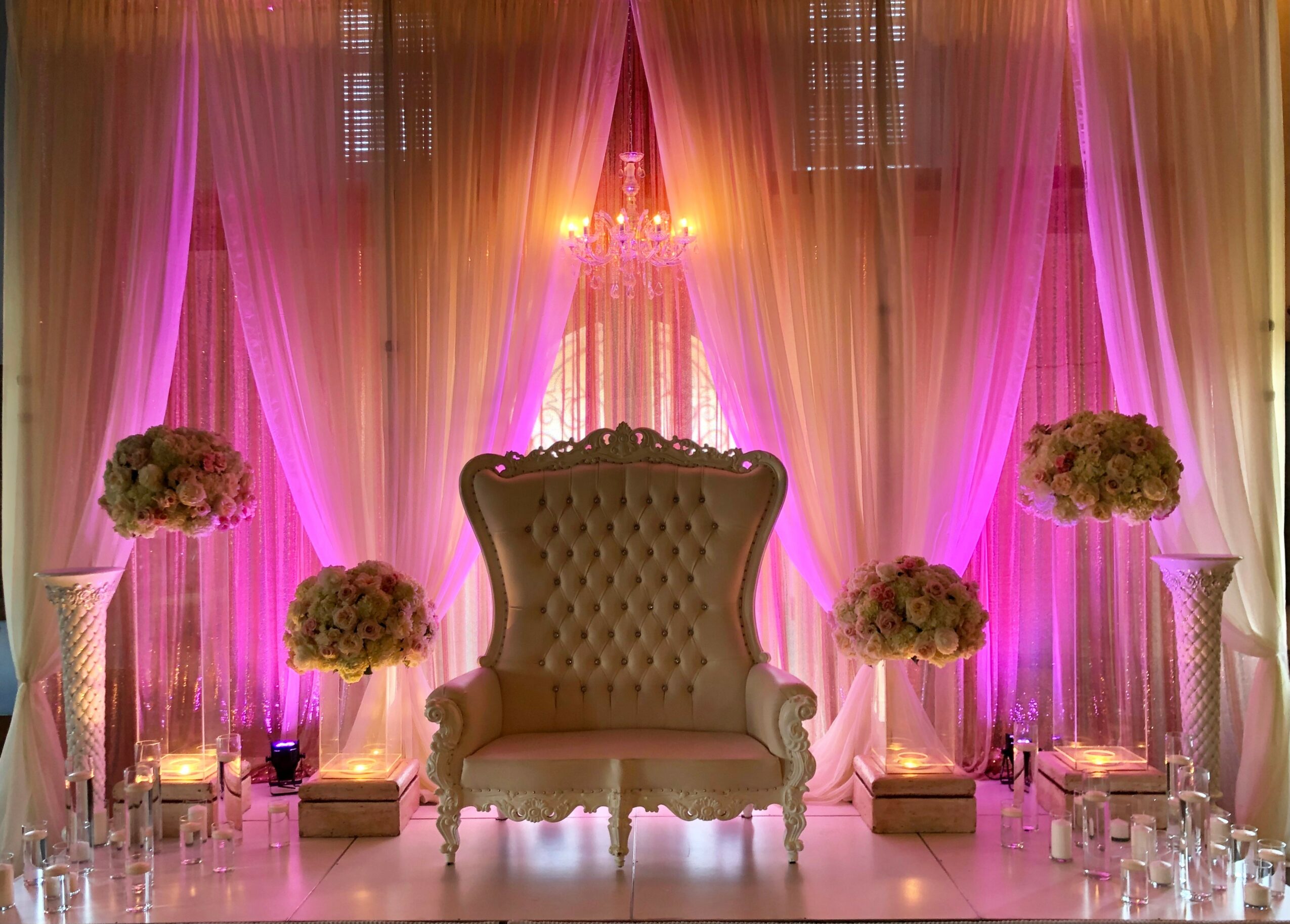 Elegant stage with a high-backed sofa, floral arrangements, pink lighting, and sheer curtains, decorated for an event or celebration.