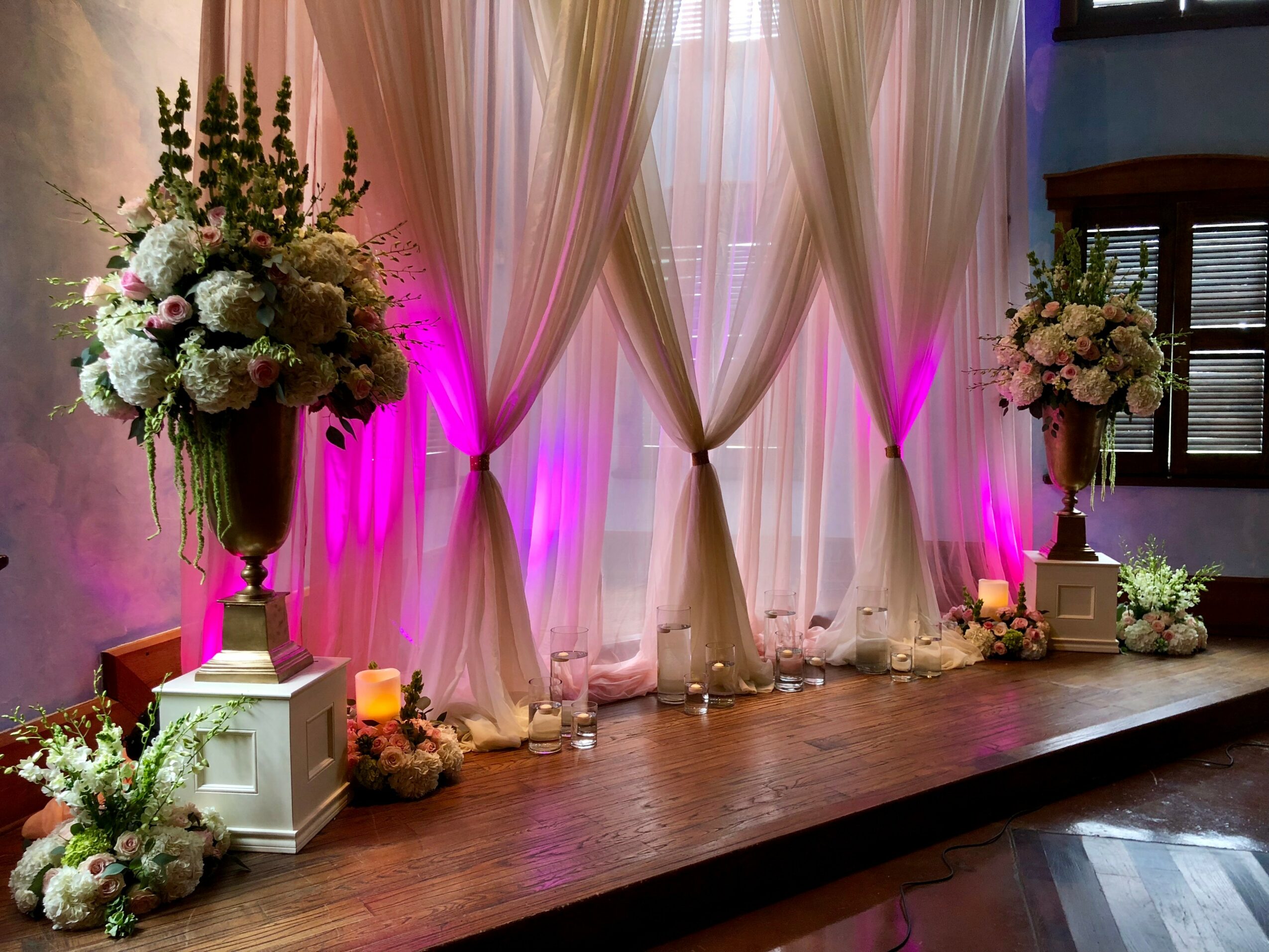 Elegant stage with floral arrangements, draped curtains, and pink lighting, set for an event or celebration.