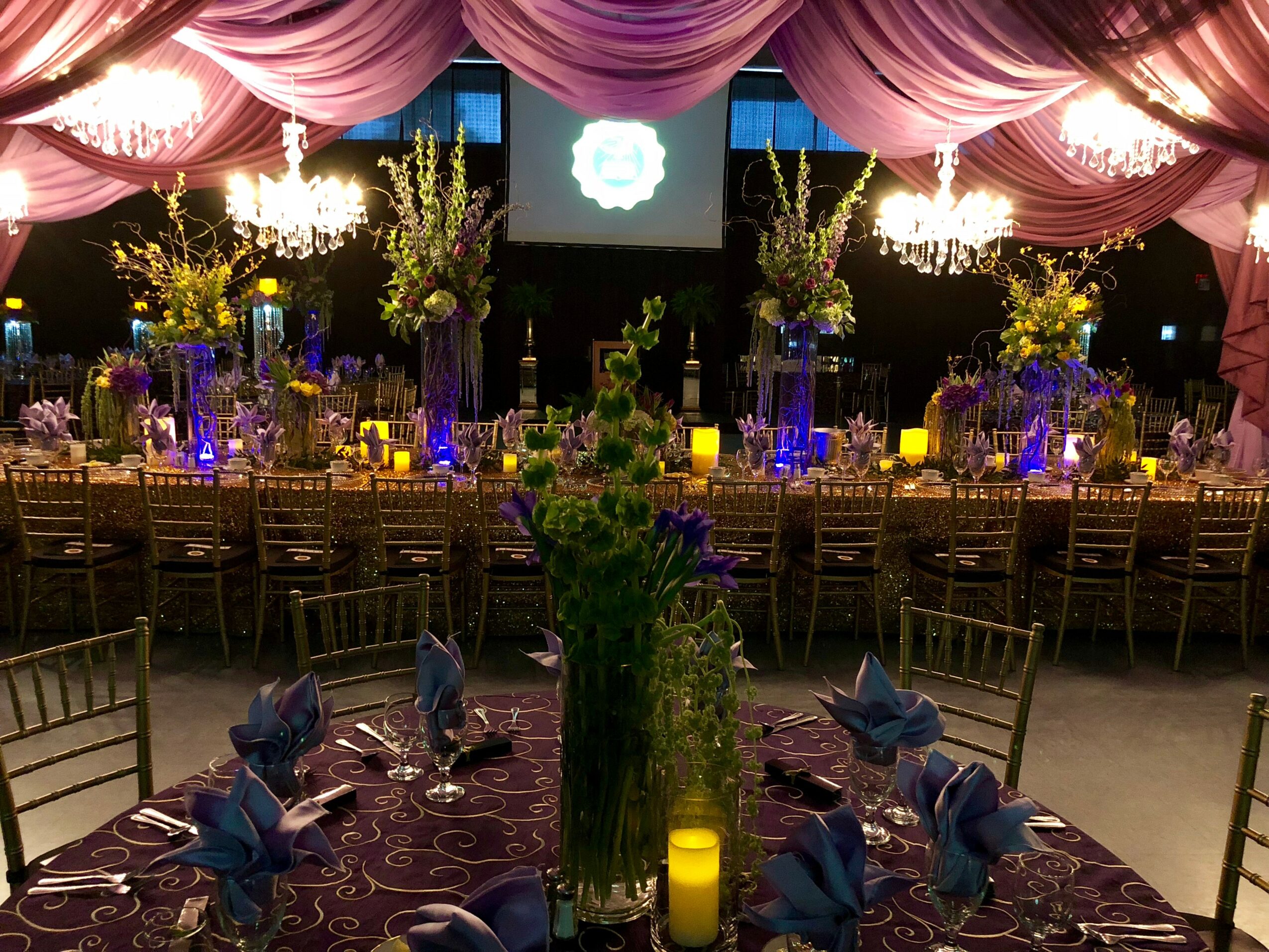 Elegant banquet hall with draped pink ceiling, chandeliers, floral centerpieces, candles, and a long decorated table.