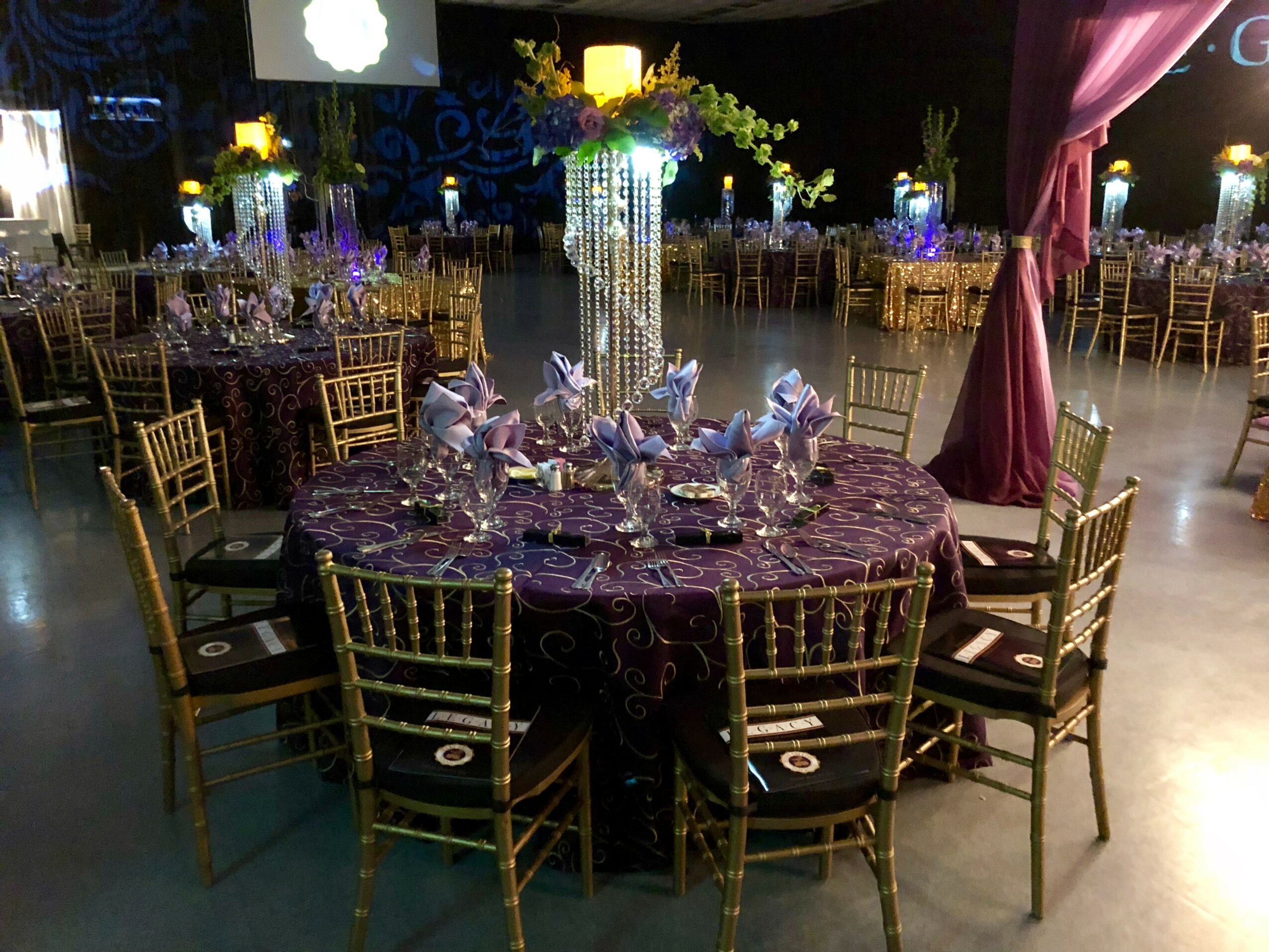 Elegant banquet hall with round table, gold chairs, floral centerpiece, and purple drapery, decorated for an event.