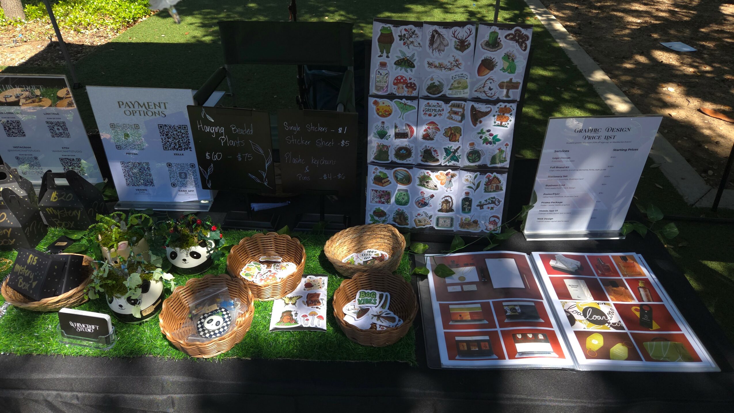 Display table with various food items, menus, and informational posters outdoors, with trees and sunlight in background.