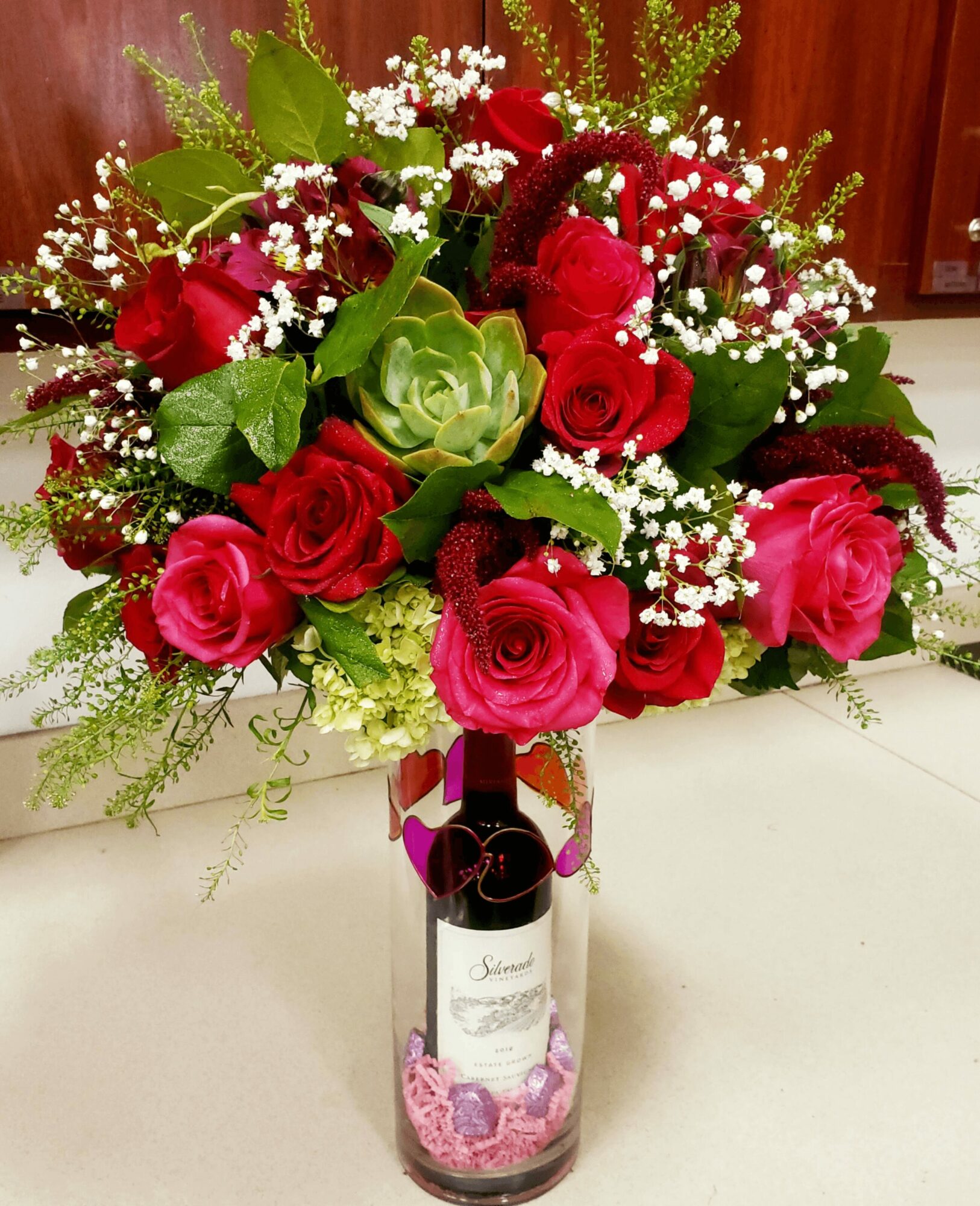 Vase with red and pink roses, white baby's breath, and green foliage on a white surface.