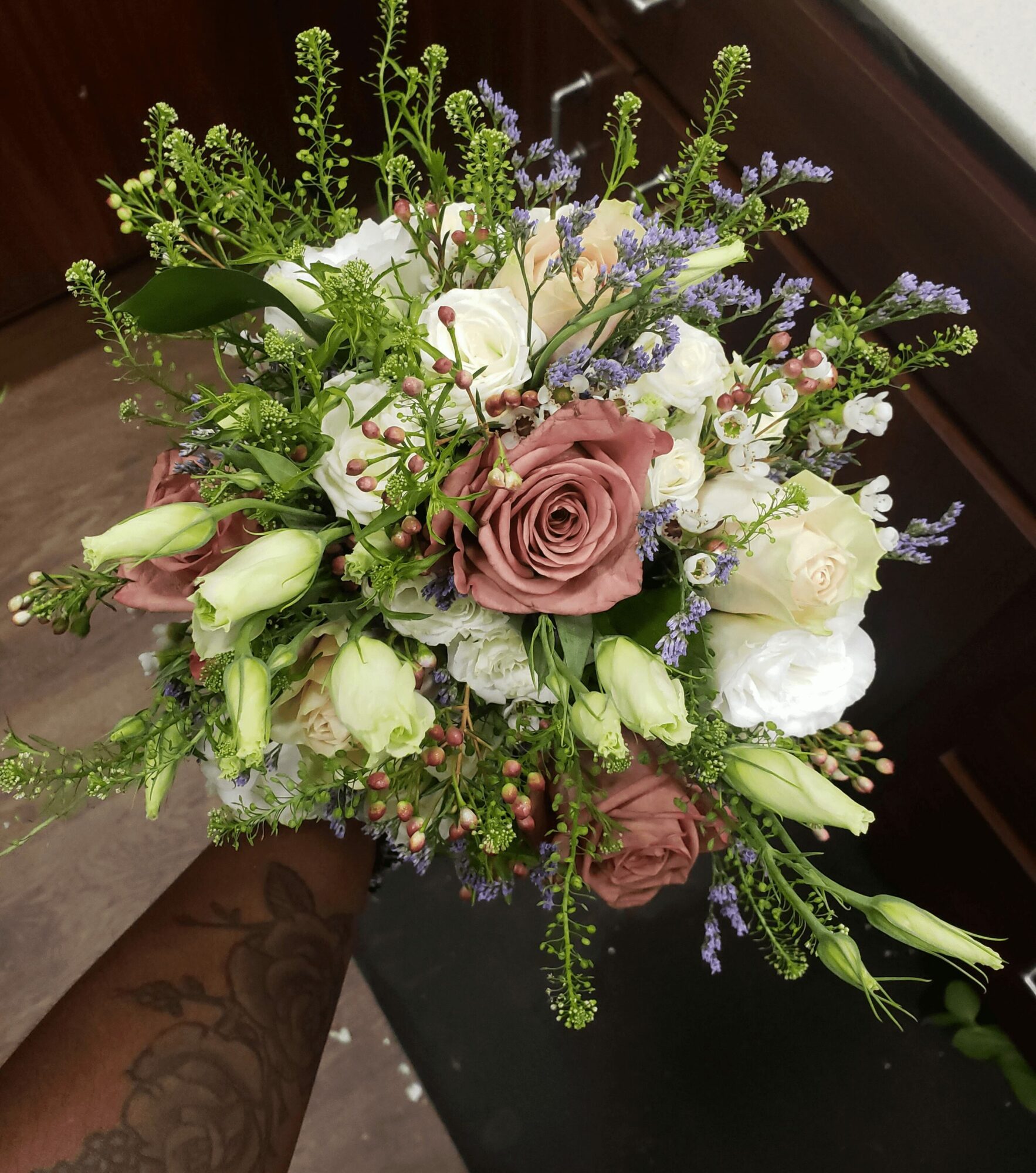 Hand holding a bouquet of pink, white, and purple flowers with green foliage, indoors near a window
