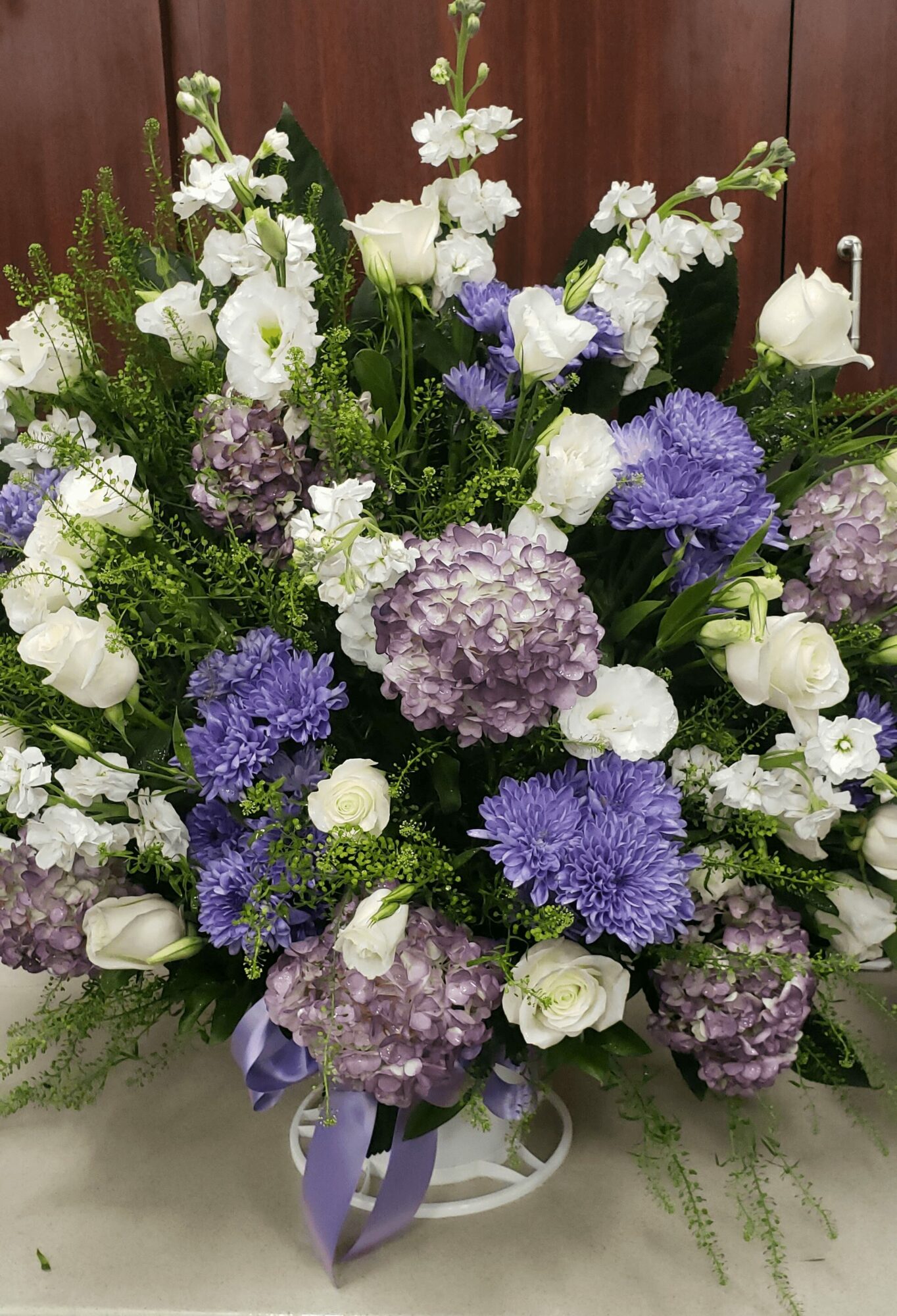 A large floral arrangement with white, purple, and lavender flowers, including roses, hydrangeas, and other blooms, in a white vase with a purple ribbon.