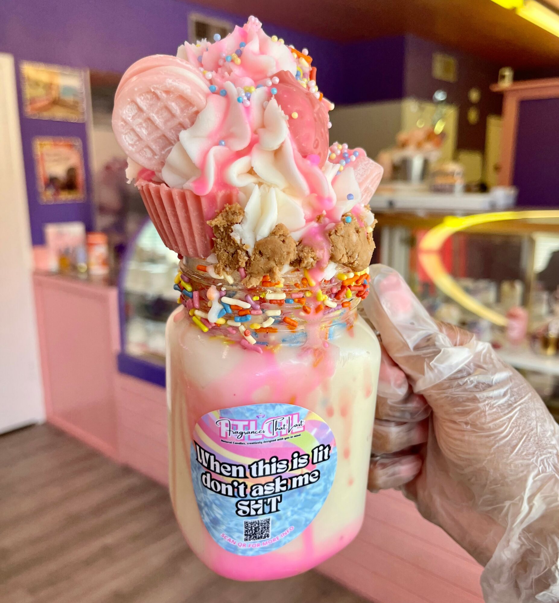 Hand holding a colorful milkshake with whipped cream, sprinkles, and candies in a pink cup, in a cafe setting.