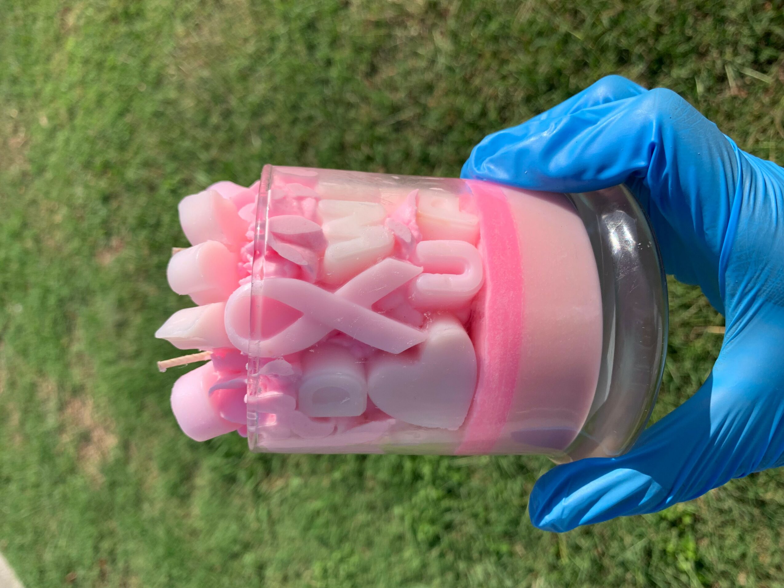 Hand in blue glove holding a glass container filled with pink and white objects, outdoors with grass background.