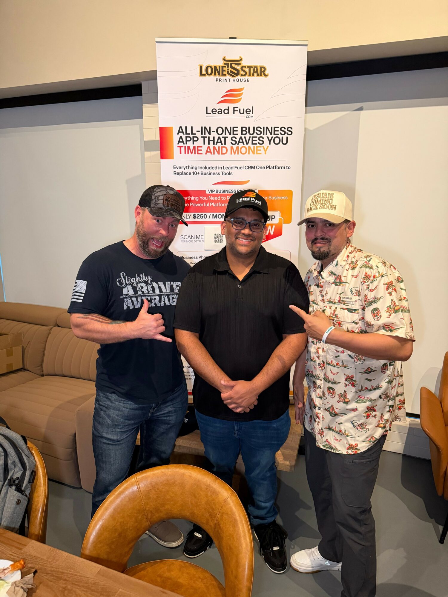 Three people standing indoors, one wearing a floral shirt and cap, another in a black shirt, and the third in a dark shirt with glasses, in front of a Louis Sar banner.