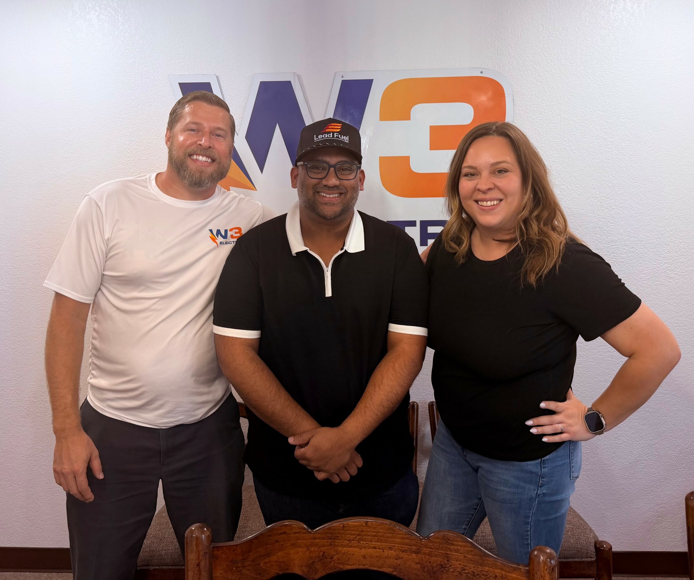 Three people standing in front of a wall with a logo, smiling, with a wooden chair in front.