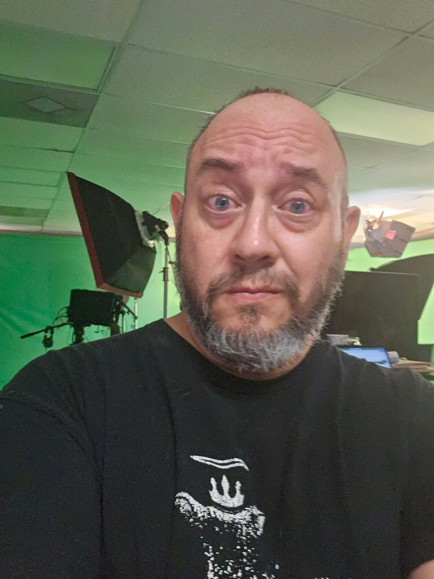 Man with a beard and bald head in a room with green and black background, wearing a black shirt.