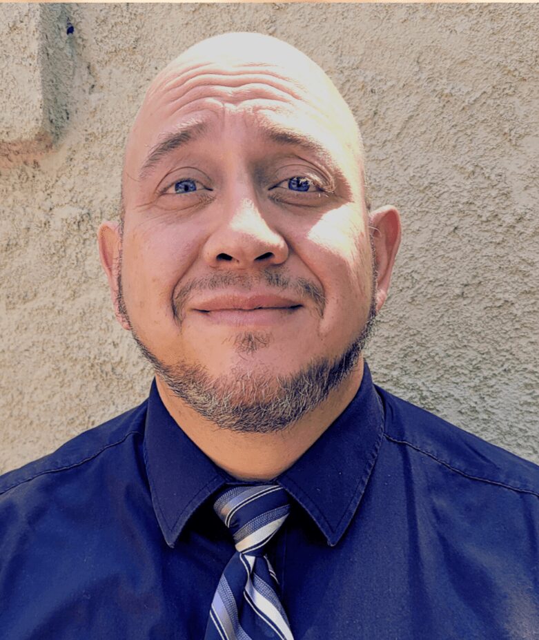 Man with a bald head, beard, wearing a dark blue shirt and striped tie, smiling slightly, standing against a textured wall.