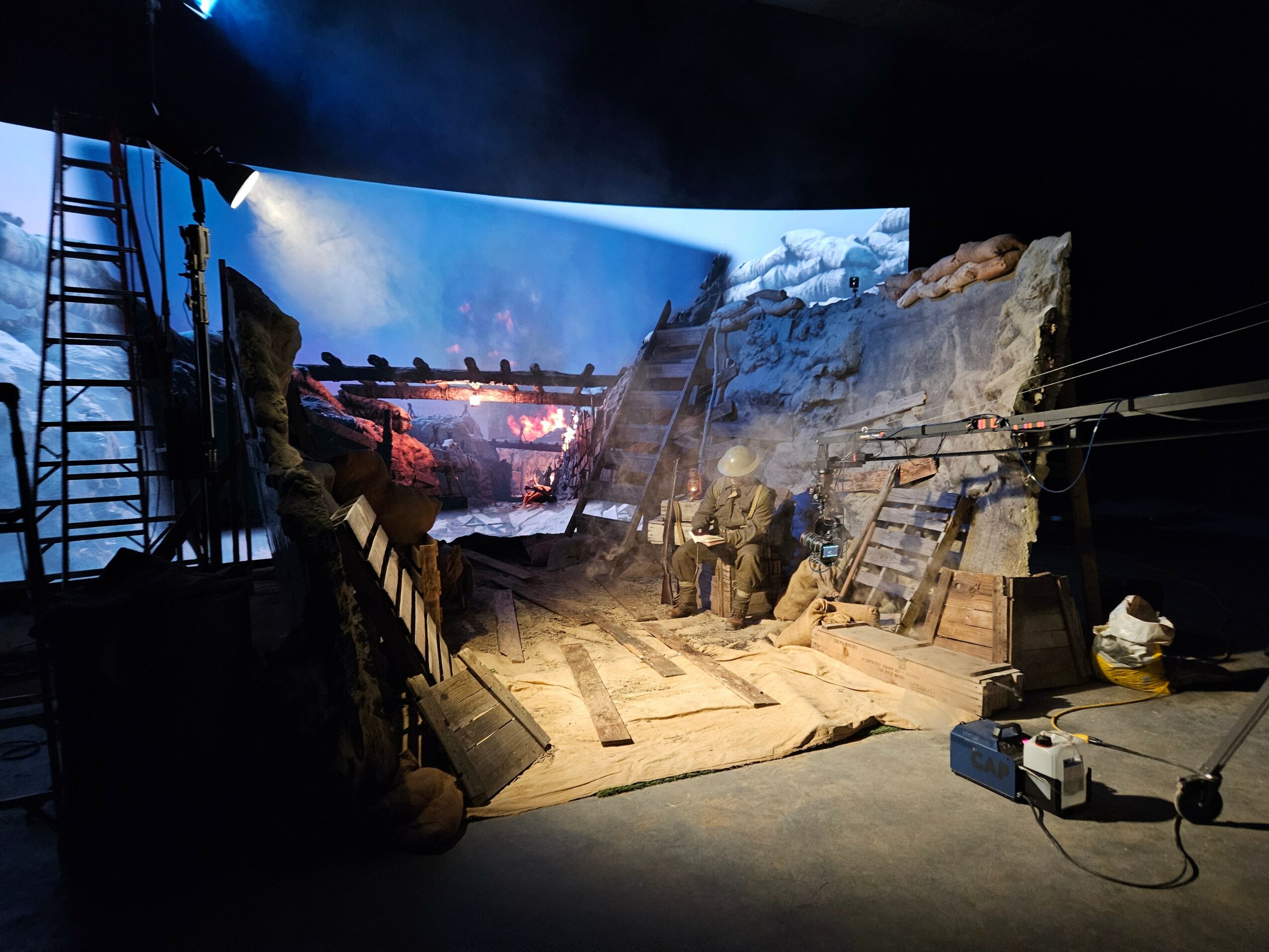 Stage set with a snowy landscape, wooden structures, and actors in winter clothing, with a backdrop of a snowy scene.