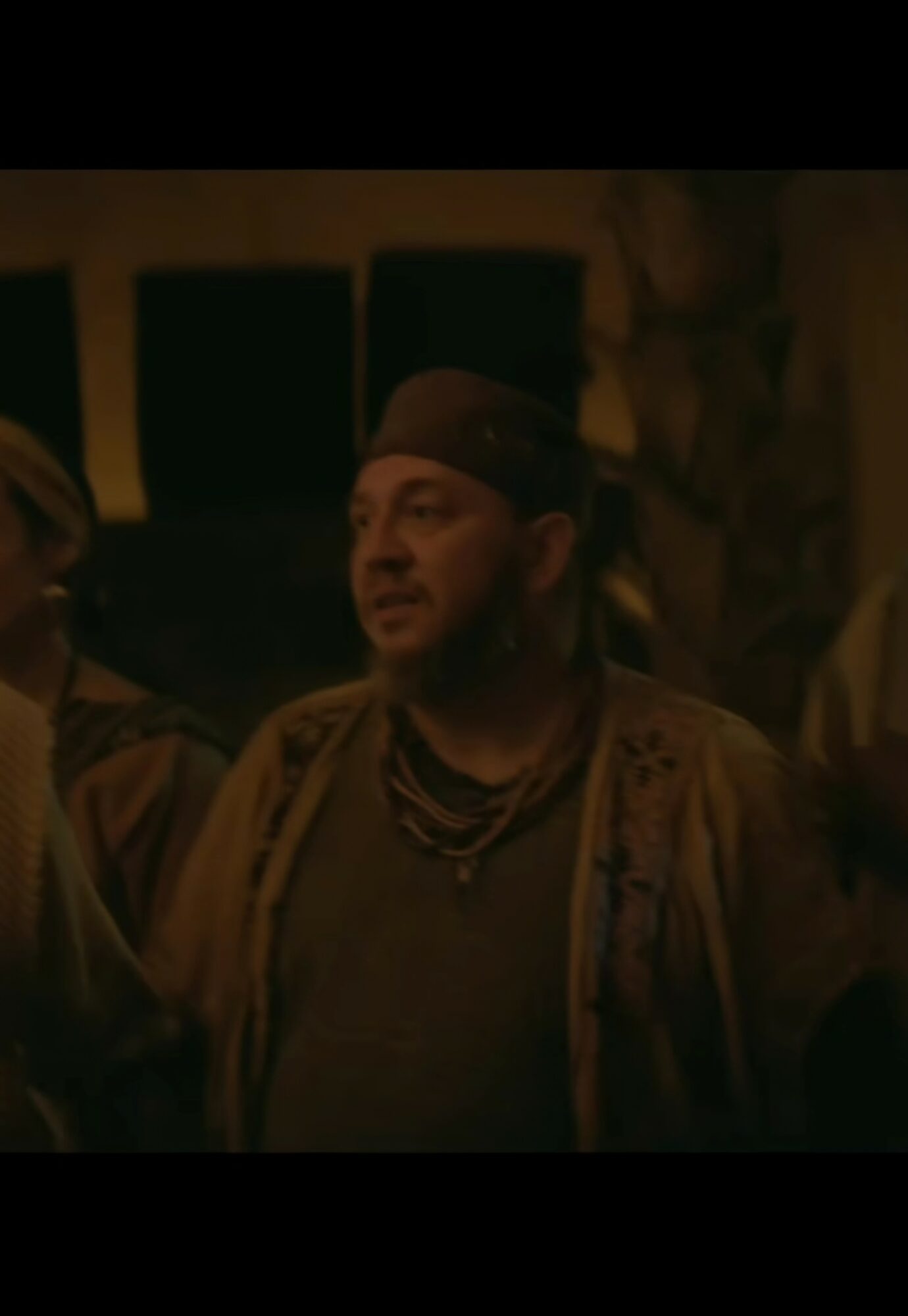 Man wearing a headscarf and layered necklaces, speaking in a dimly lit setting with dark background.