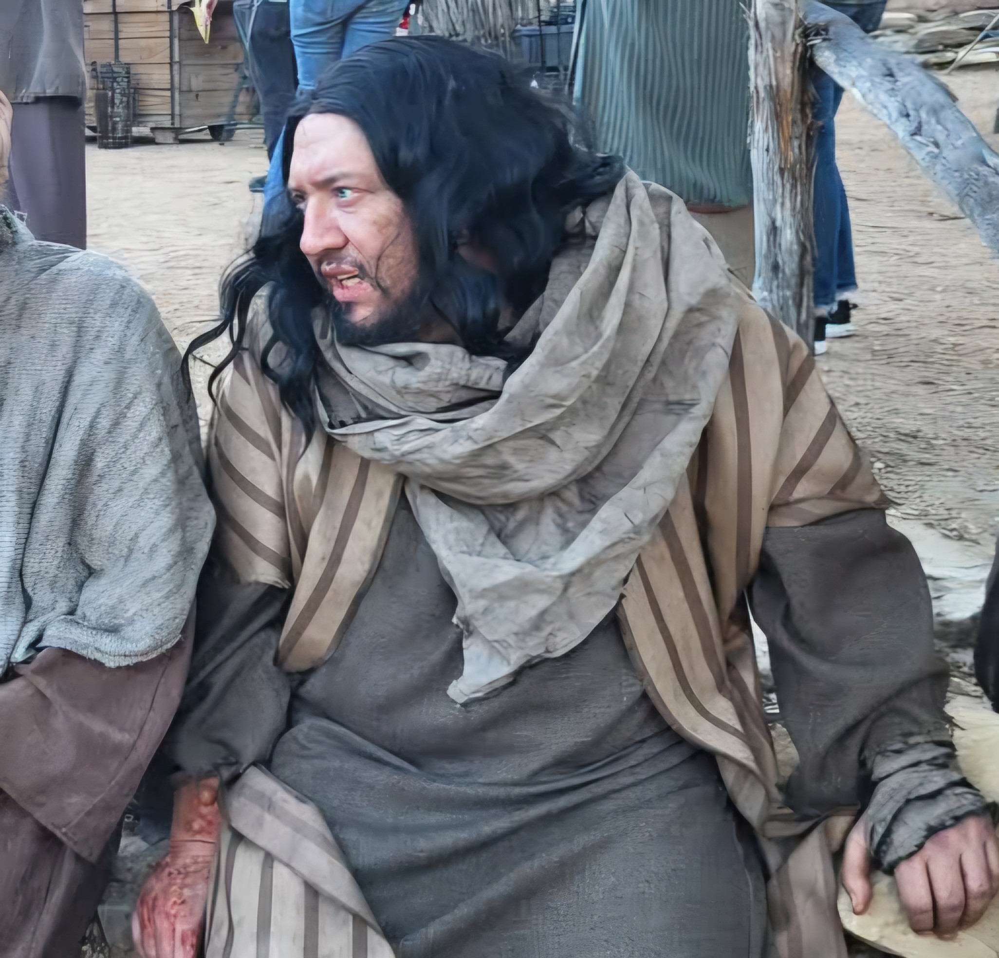 Person with long black hair, wearing a striped cloak and large scarf, sitting outdoors with a wooden structure nearby.