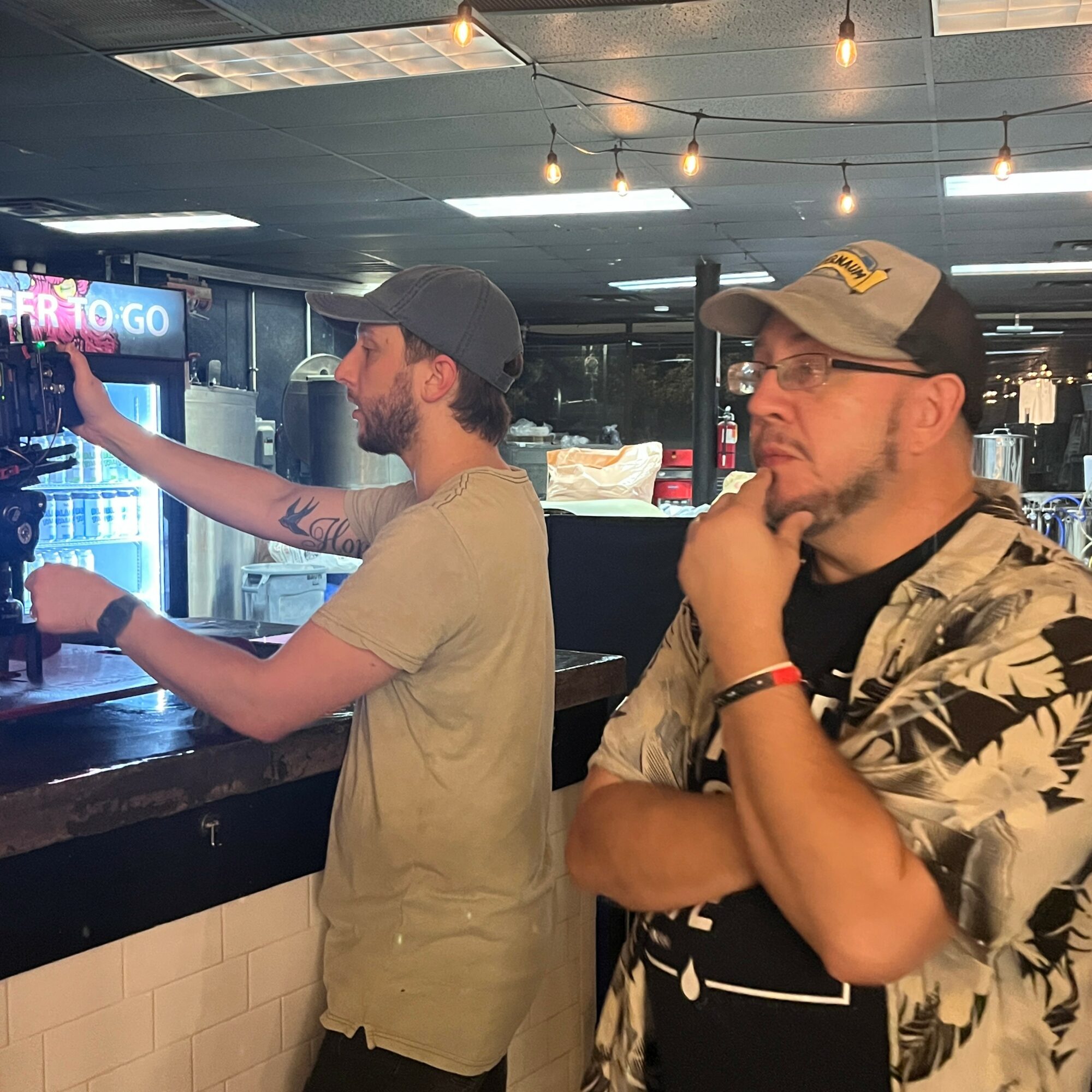 Two men standing at a bar, one reaching for a drink, the other with hand on chin, in a dimly lit setting.