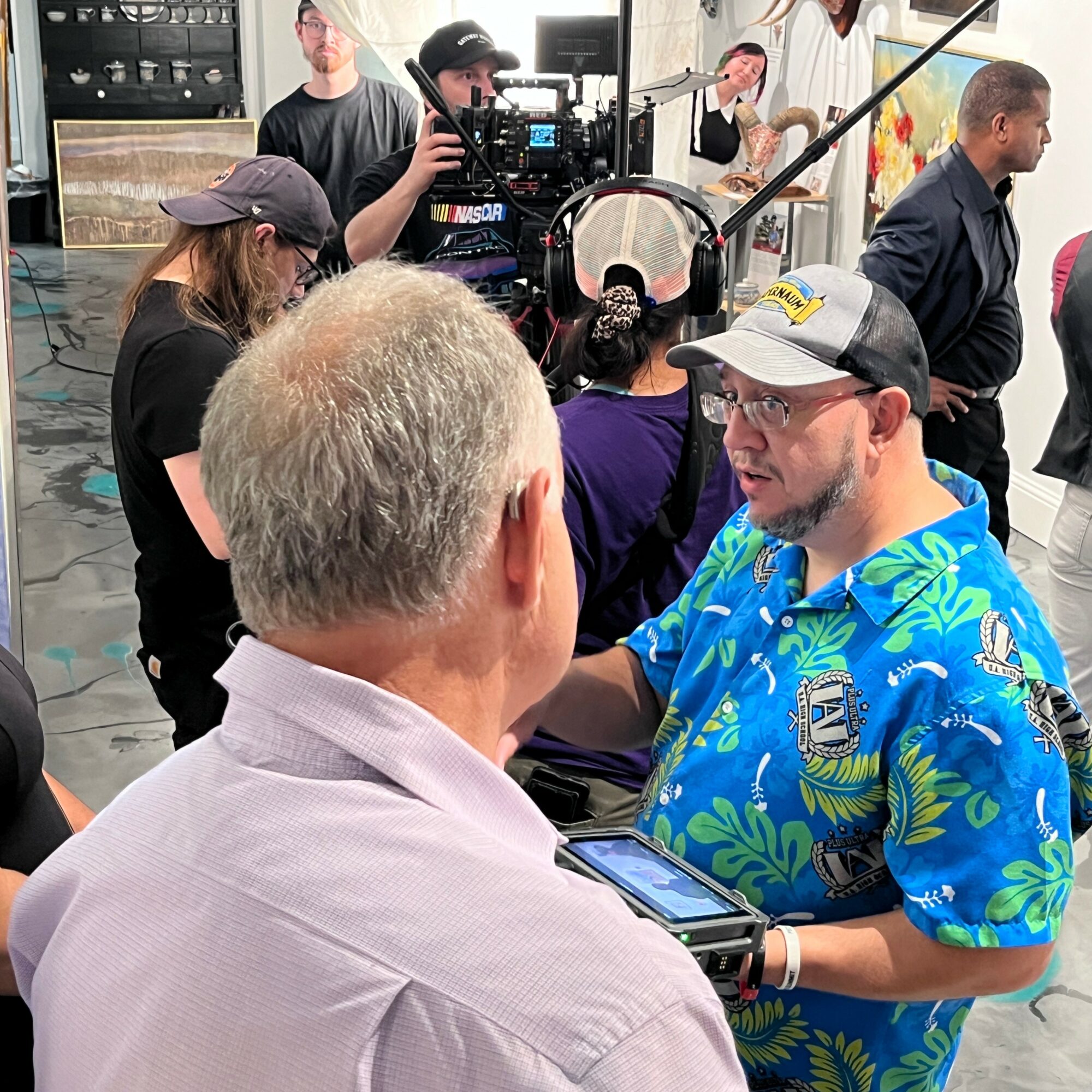 Two men engaged in conversation, one with gray hair and the other wearing a colorful Hawaiian shirt and cap, in a busy indoor setting.