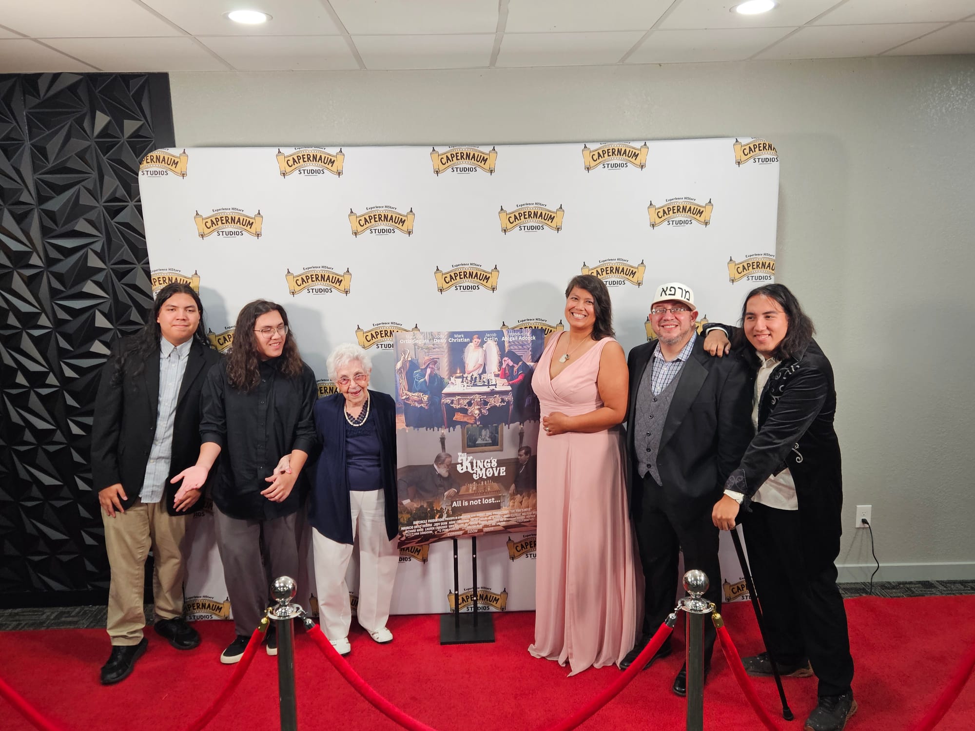 Group of seven people standing on a red carpet in front of a backdrop with logos, posing for a photo.