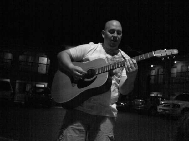Person playing guitar on stage in a dark indoor setting, with visible balcony and parked cars in background.