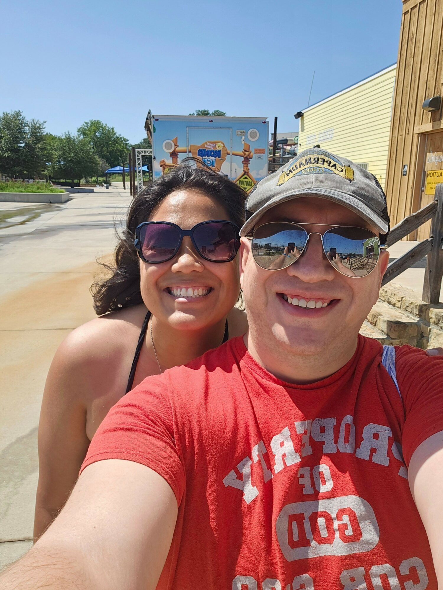 Two women smiling outdoors, one wearing sunglasses and a white cap, the other wearing sunglasses, with a blue sky and a sign in the background.