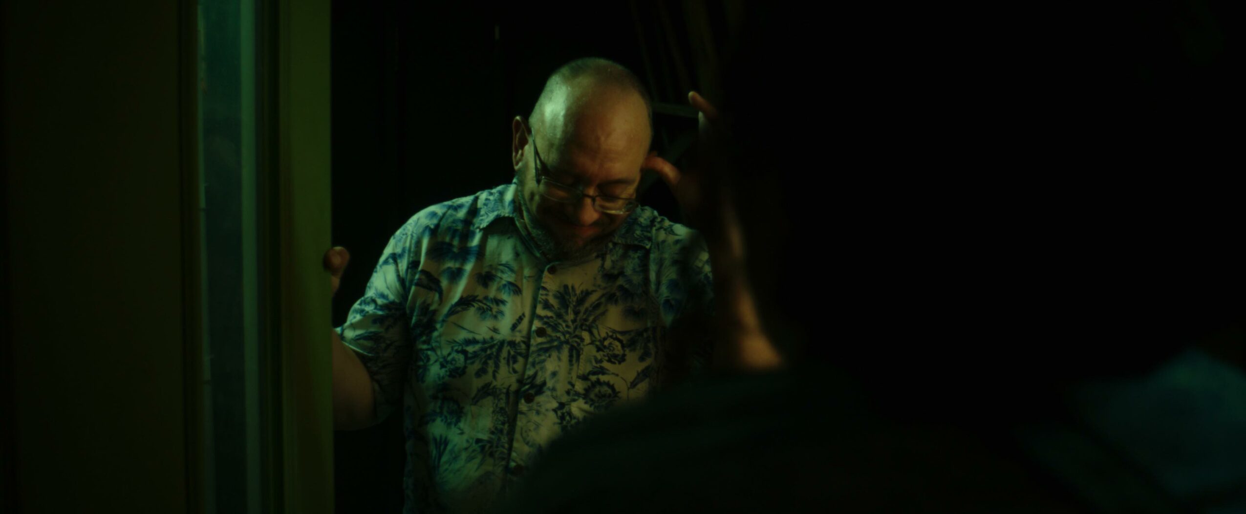 Person with glasses and patterned shirt looking down, partially visible in a dark setting.
