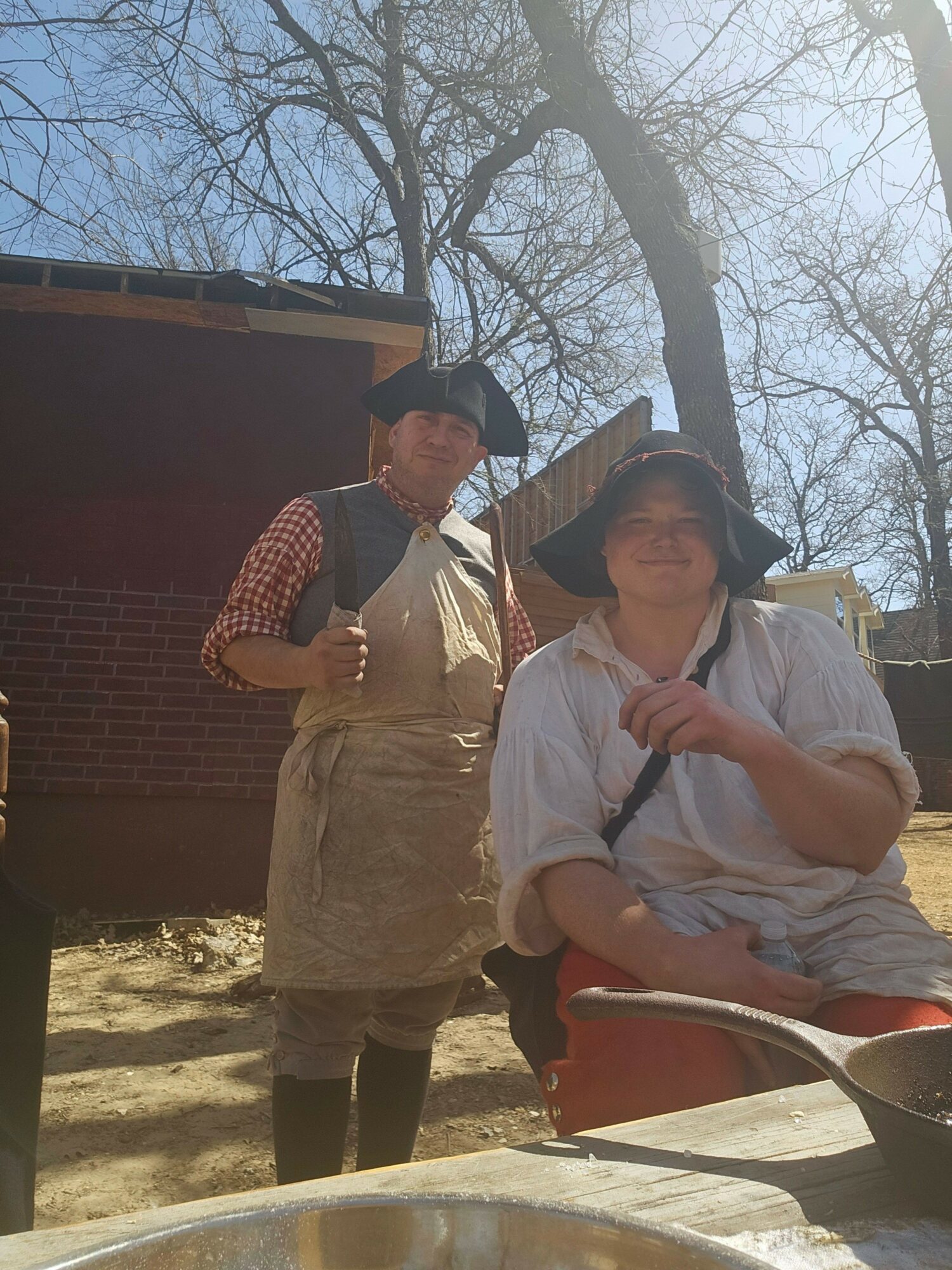Two people sitting outdoors, one in a white shirt and apron, the other in a checkered shirt and apron, with trees in background.