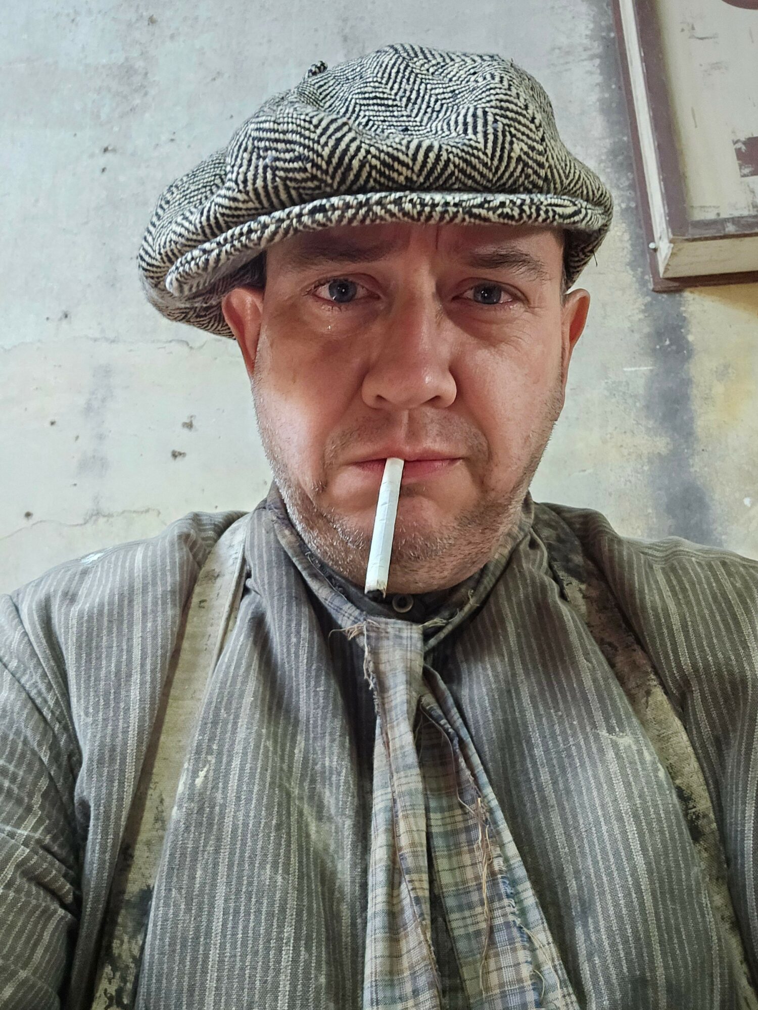 Man wearing a patterned cap and striped shirt, smoking a cigarette, with a concrete wall background.
