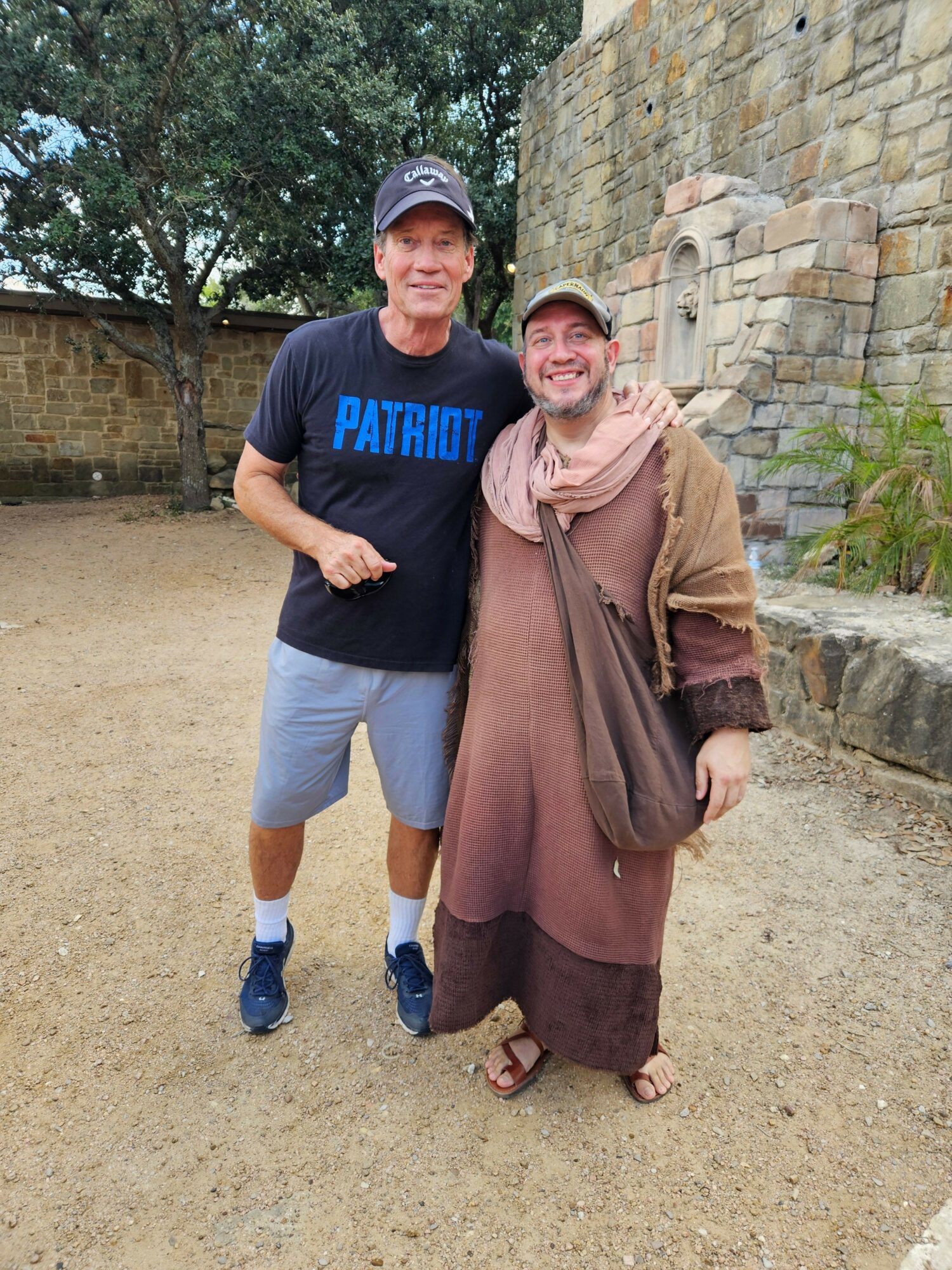 Two people standing outdoors on a dirt path, one woman in a long dress and headscarf, one man in a T-shirt and shorts.