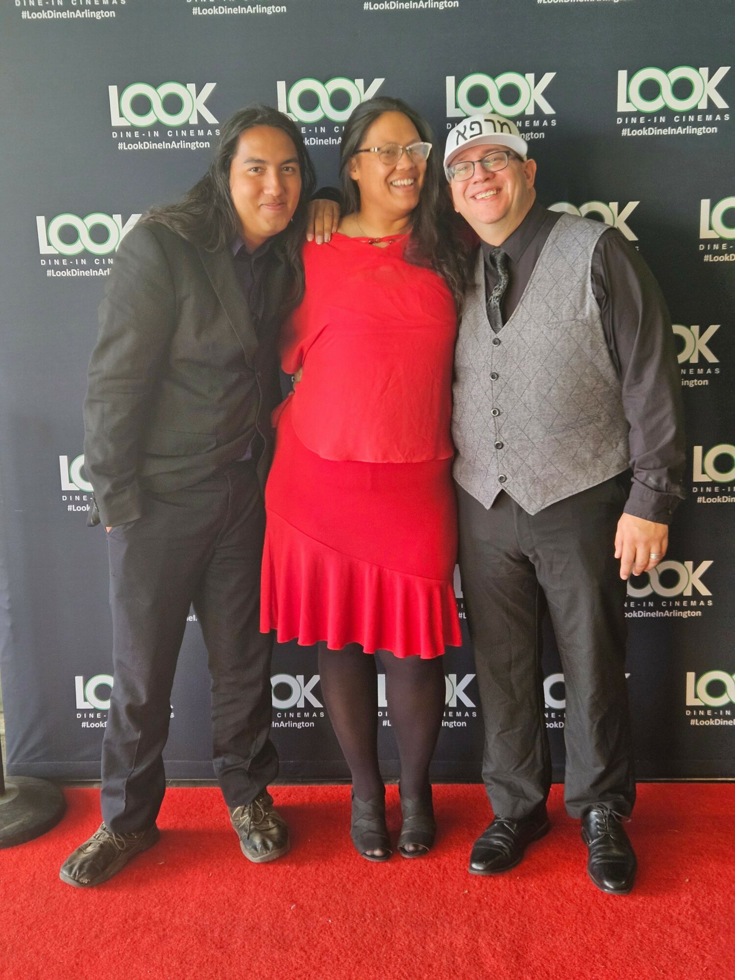 Three people standing on a red carpet in front of a black backdrop with white text, smiling at the camera.