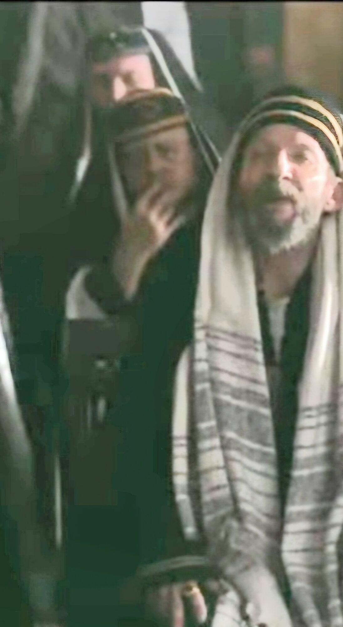 Two men wearing traditional head coverings, one with a striped scarf, standing close together, one with hand near face.