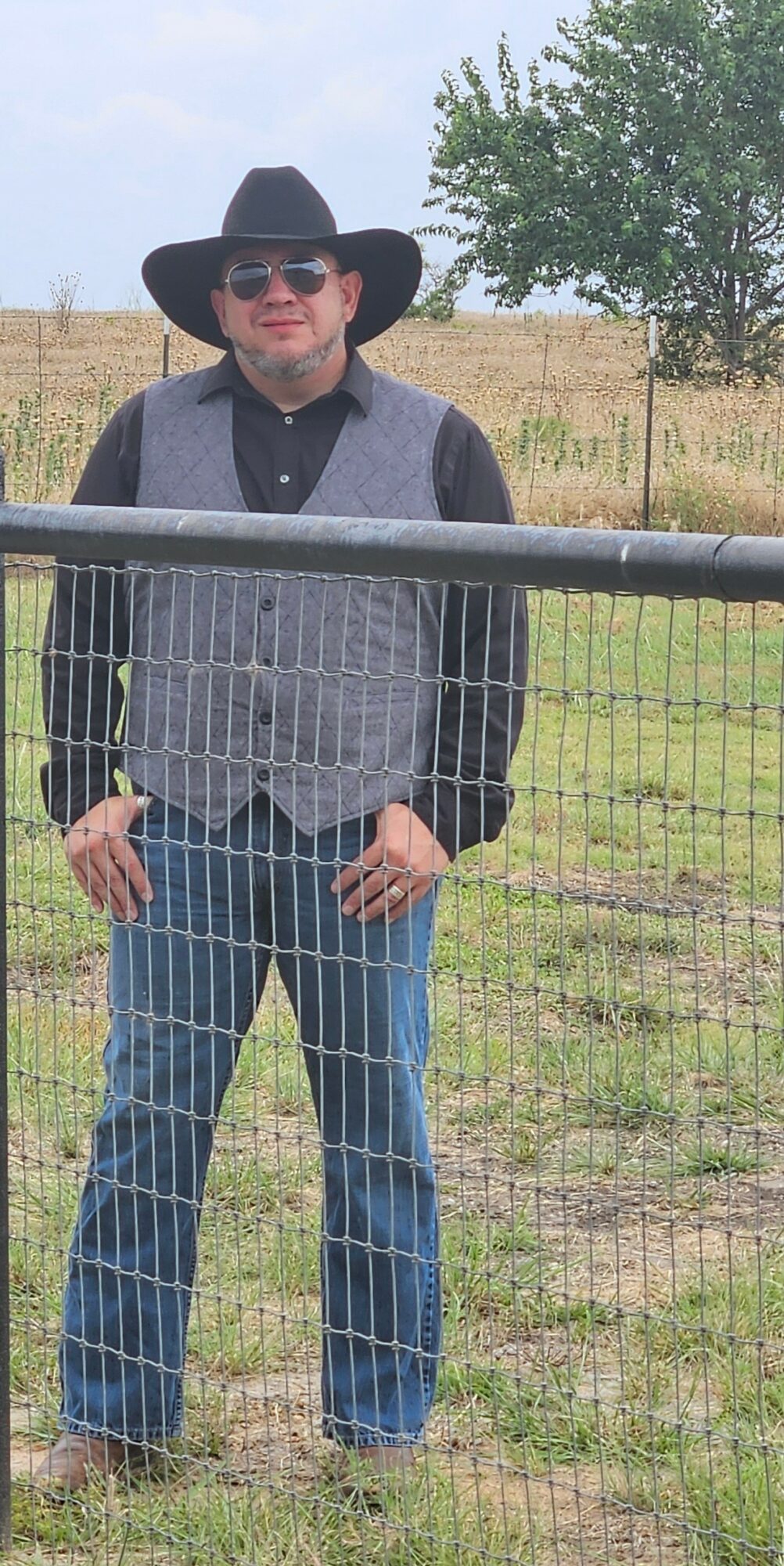 Person wearing a large black hat, sunglasses, gray vest, black shirt, and jeans standing outdoors behind a wire fence.