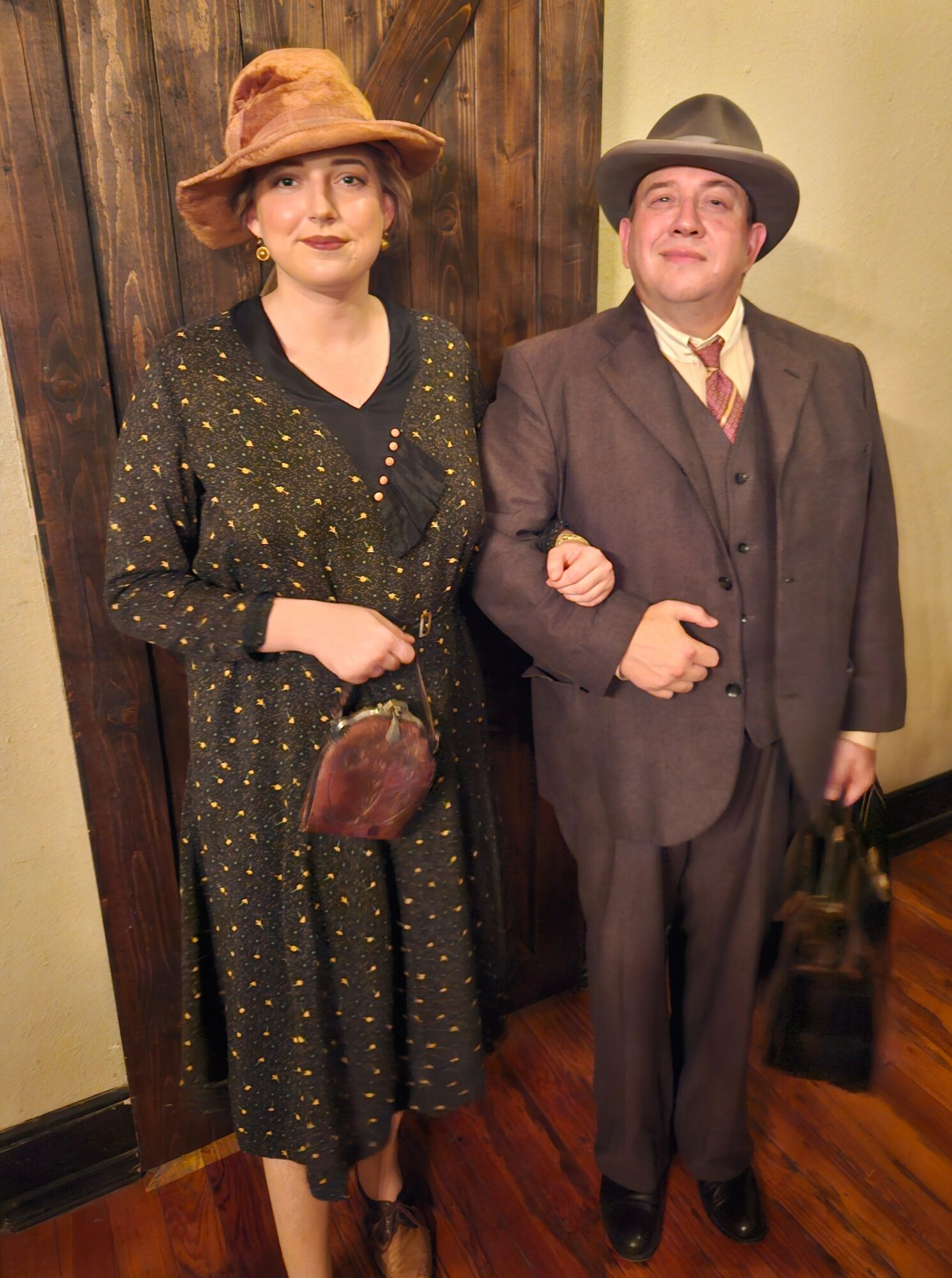 Two people dressed in vintage clothing, standing indoors against a wooden wall, with one person holding a bag.