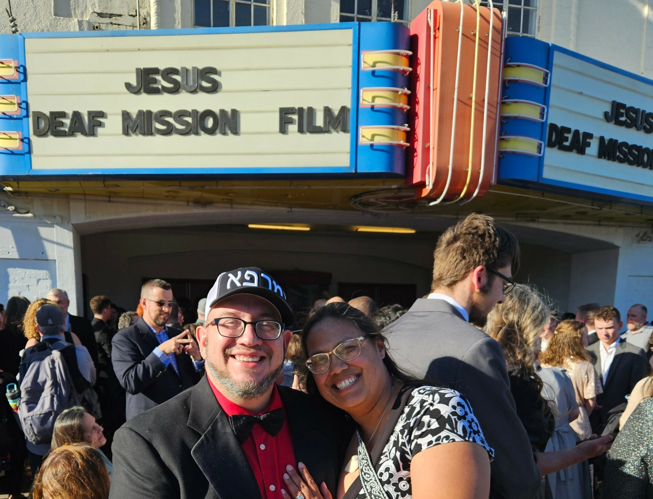 Two people smiling in front of a theater marquee with the words 'JESUS DEAF MISSION FILM' visible, surrounded by a crowd.