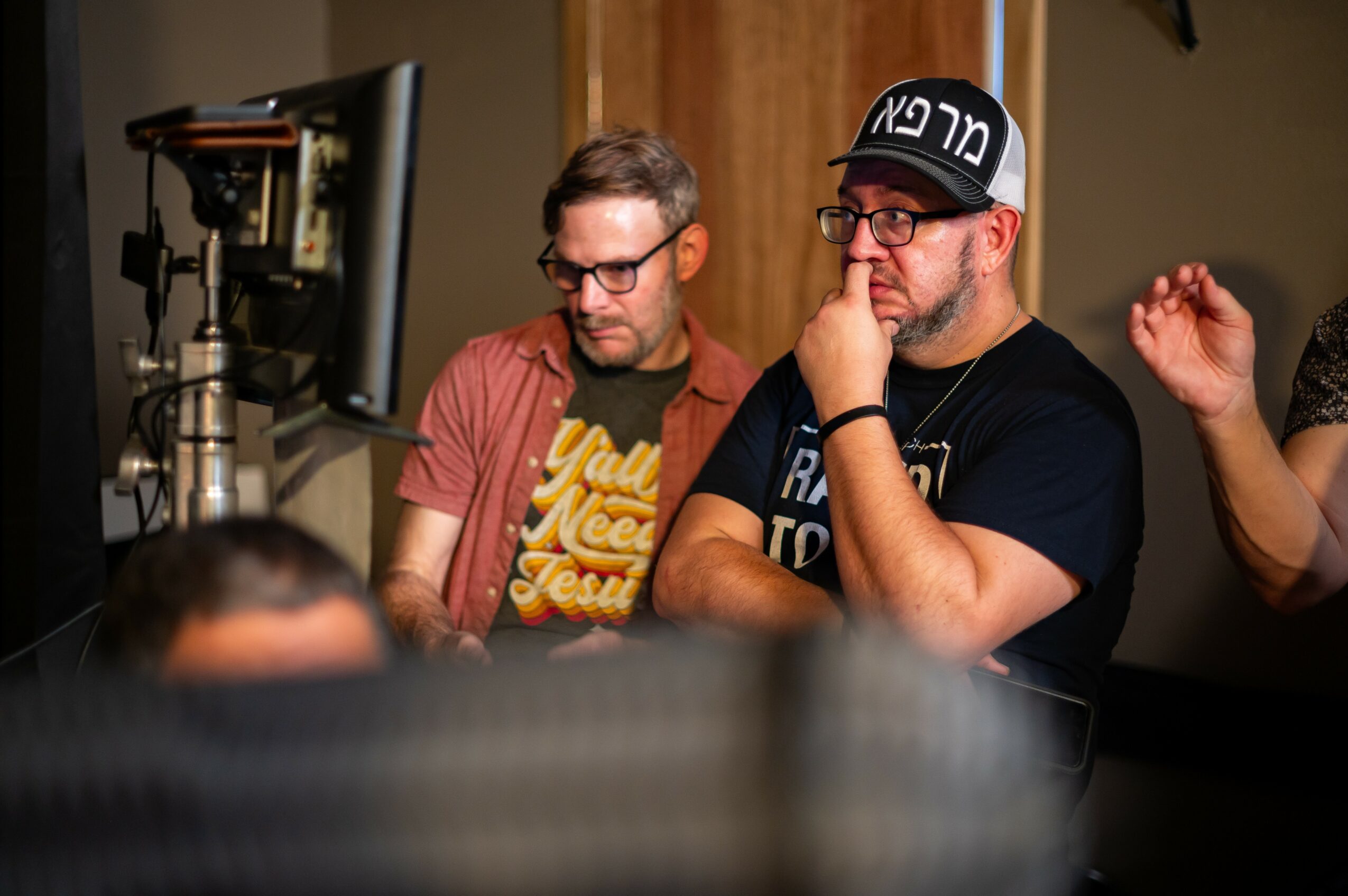 Two men sit at a desk looking at a computer monitor, one wearing glasses and a red shirt, the other wearing a cap and black shirt.