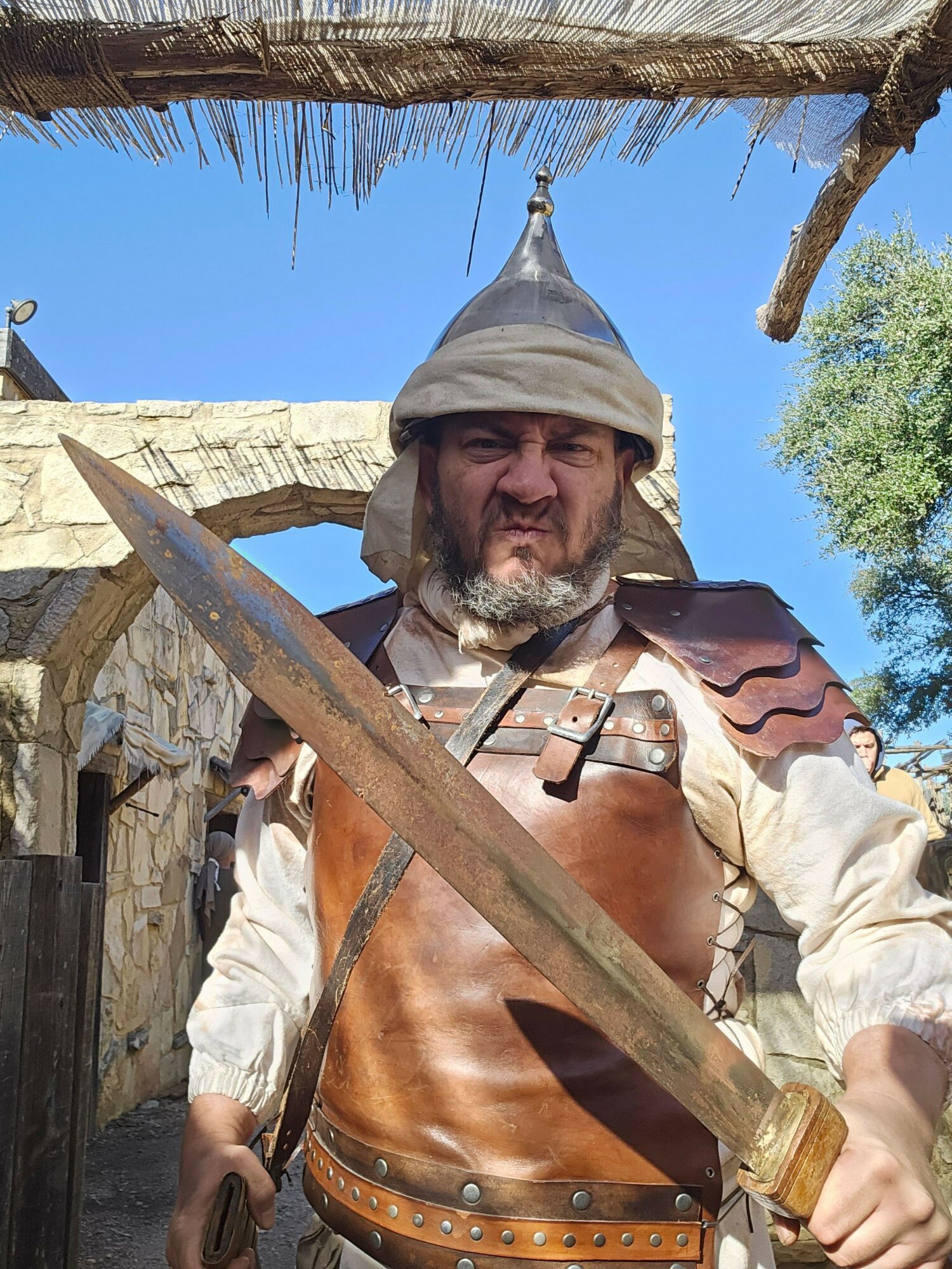 Man dressed in medieval costume holding a sword outdoors with stone structures and trees in background.