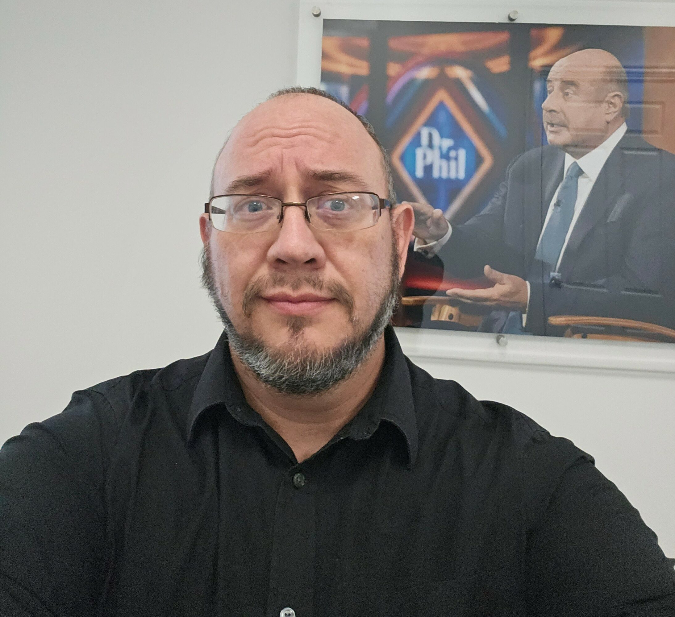 Man with glasses and beard taking selfie in front of a Dr. Phil show poster
