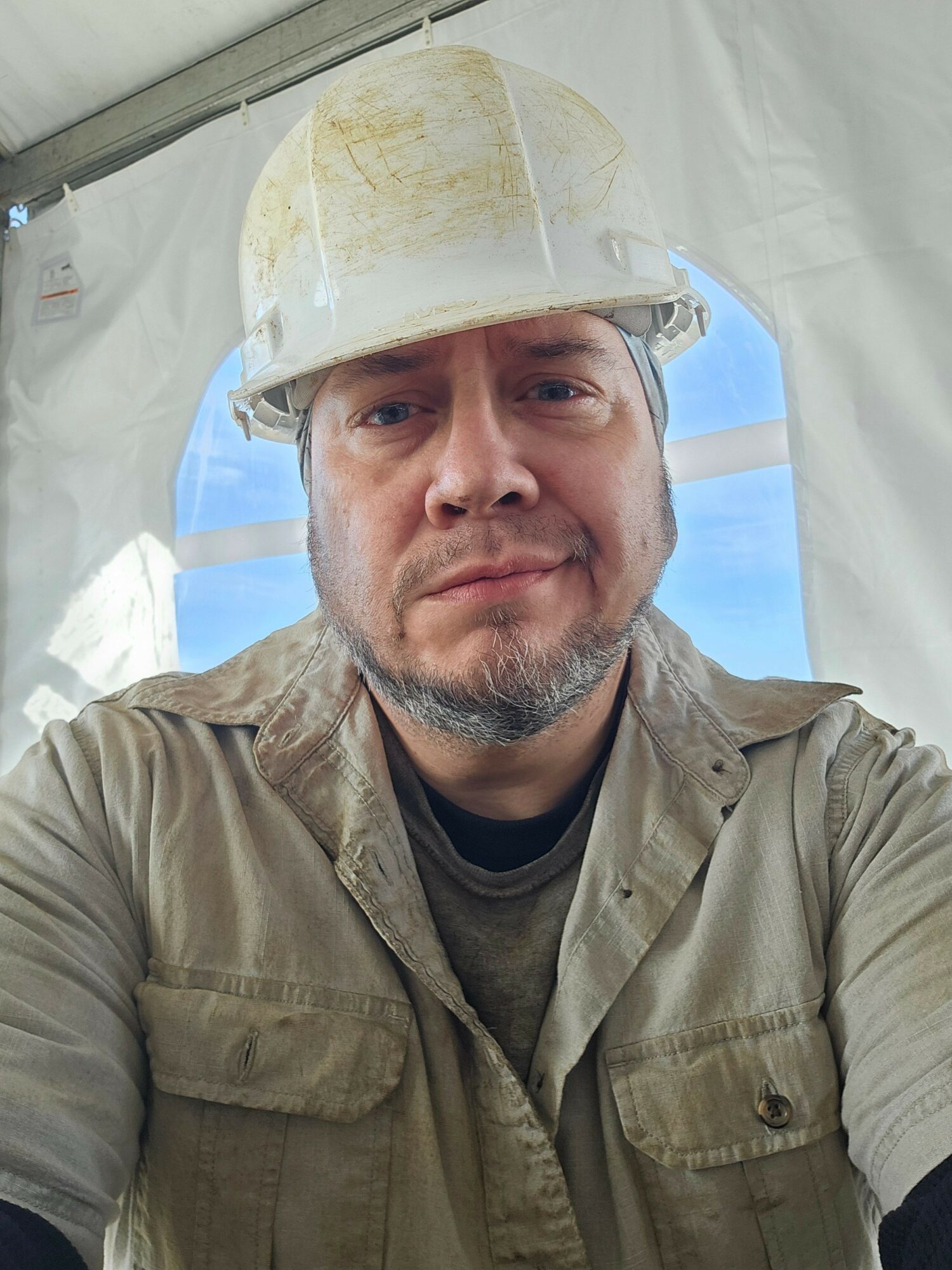 Man wearing a white hard hat and beige jacket, smiling inside a tent with a blue sky background.