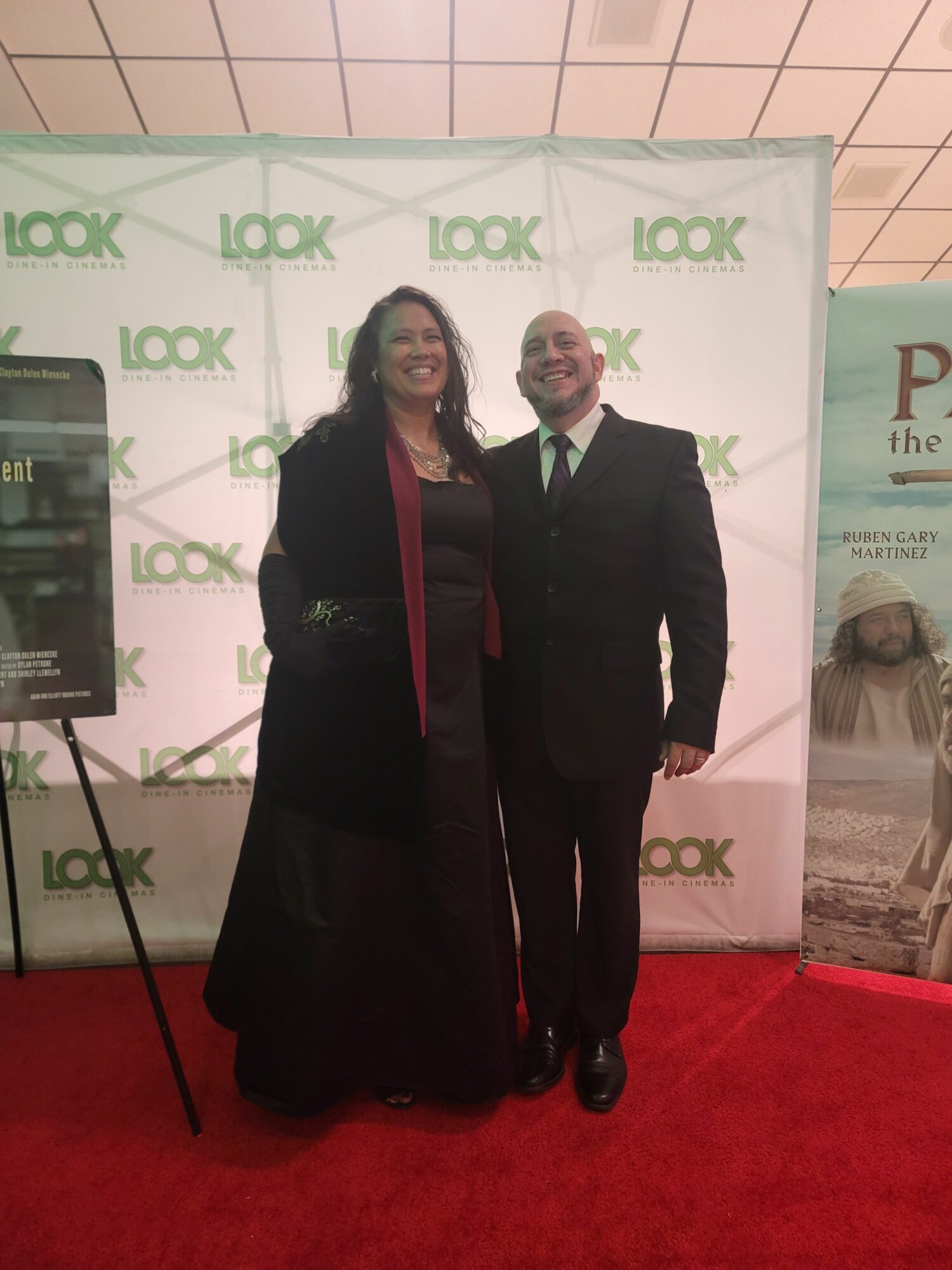 Two people standing in front of a backdrop with green and white logo, one in a suit and the other in a coat, on a red carpet.