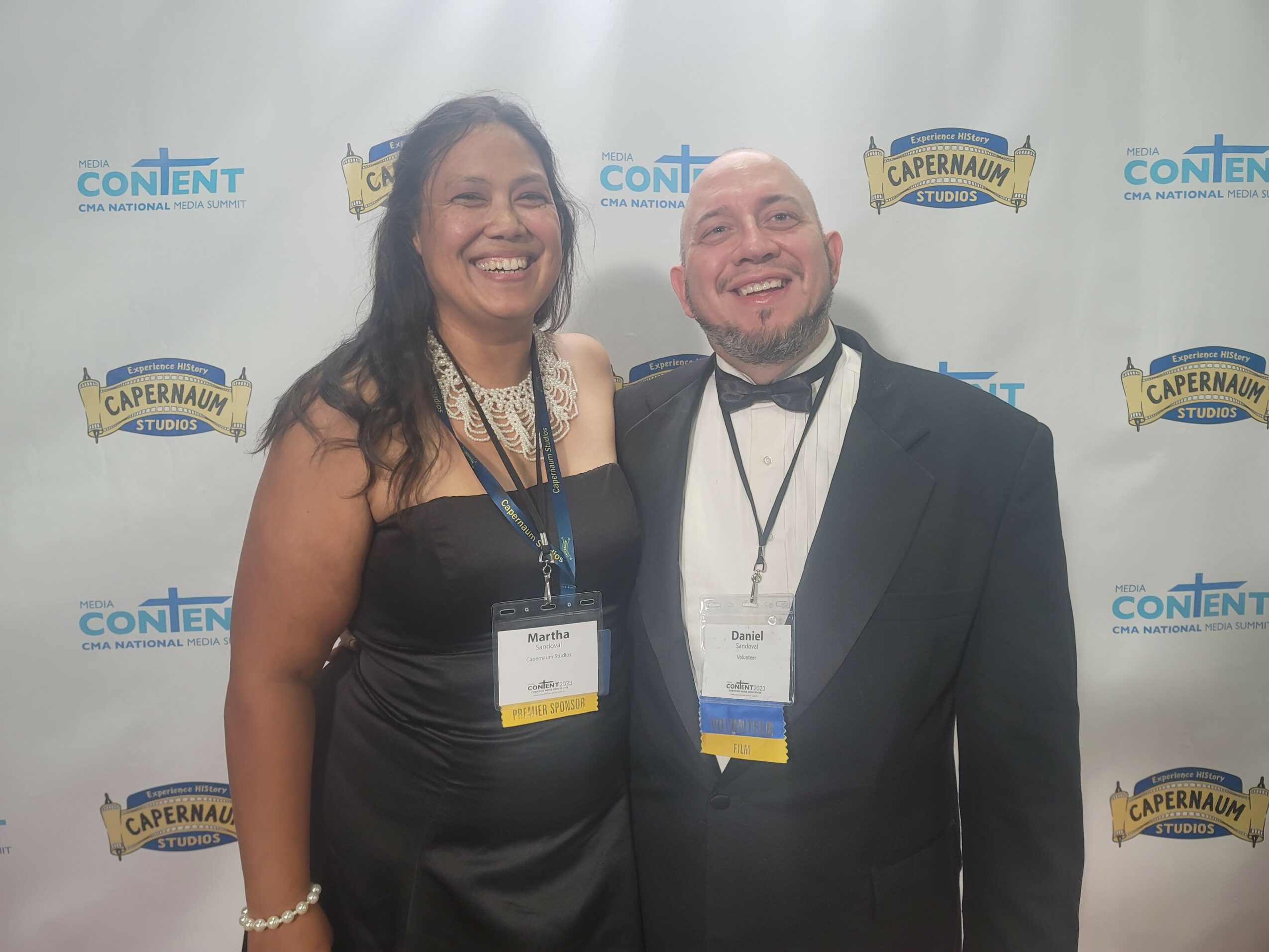 Two people smiling, one woman in a black dress and one man in a tuxedo, standing in front of a backdrop with logos.