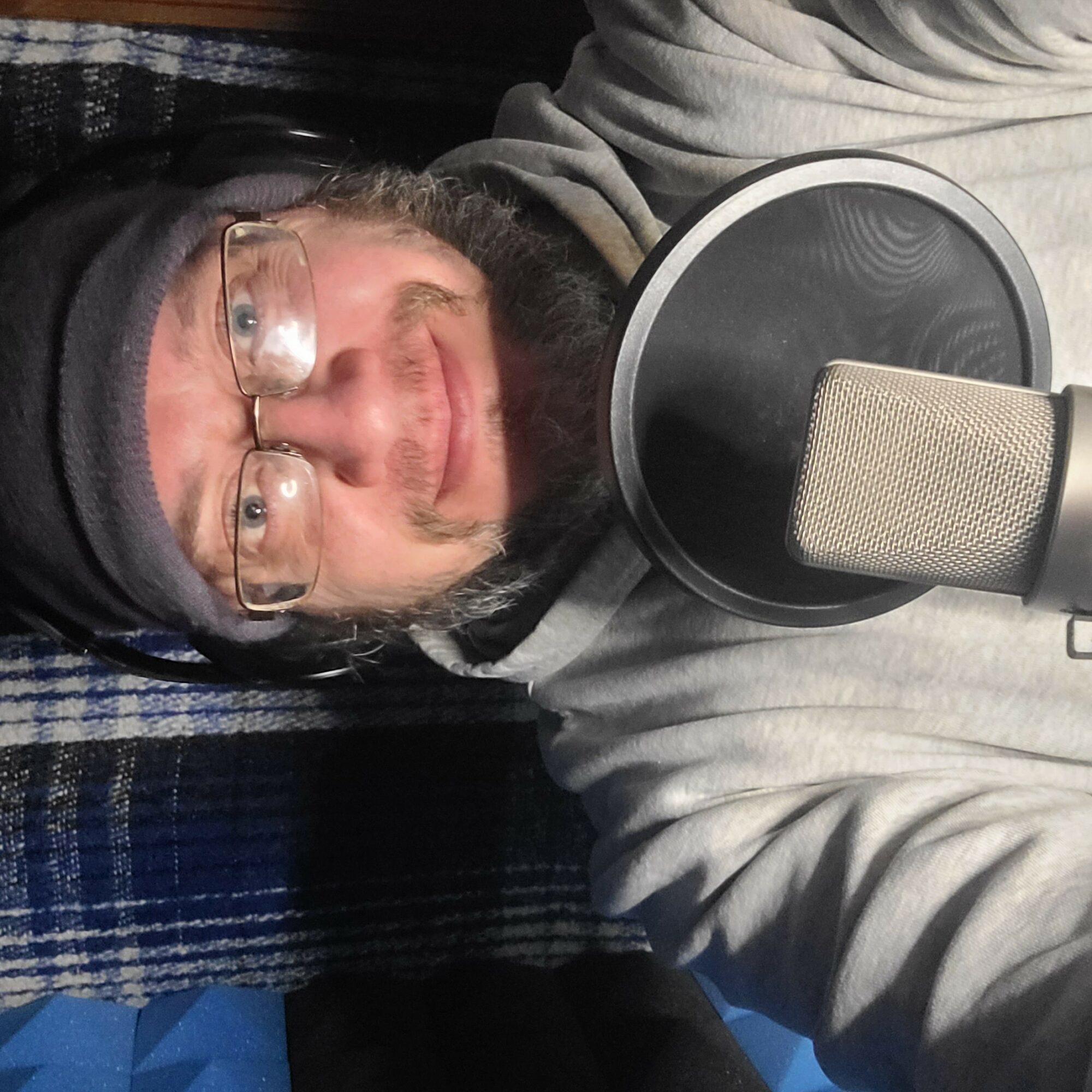 Person with glasses and a beard recording into a microphone, lying down on a striped surface.