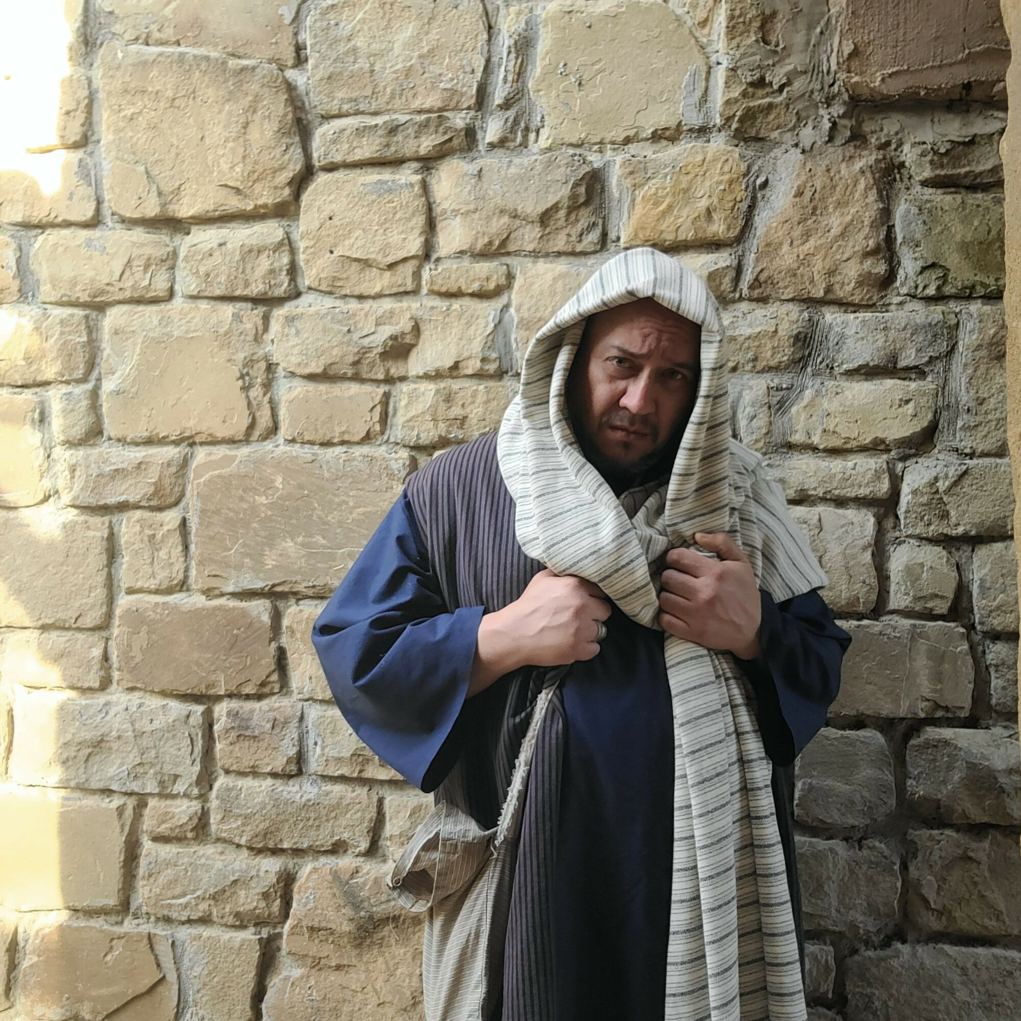 Person wearing a hooded cloak standing against a stone wall, holding the cloak with both hands.
