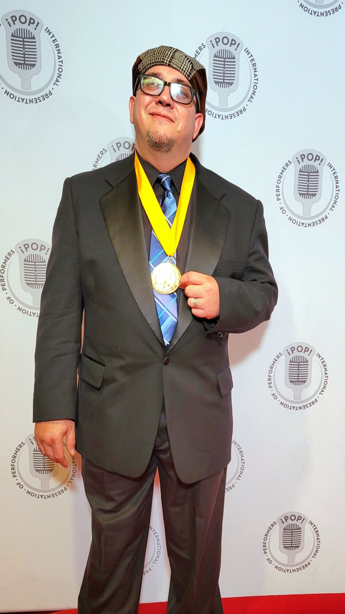 Person in a suit with glasses and a medal, standing in front of a backdrop with logos, smiling.