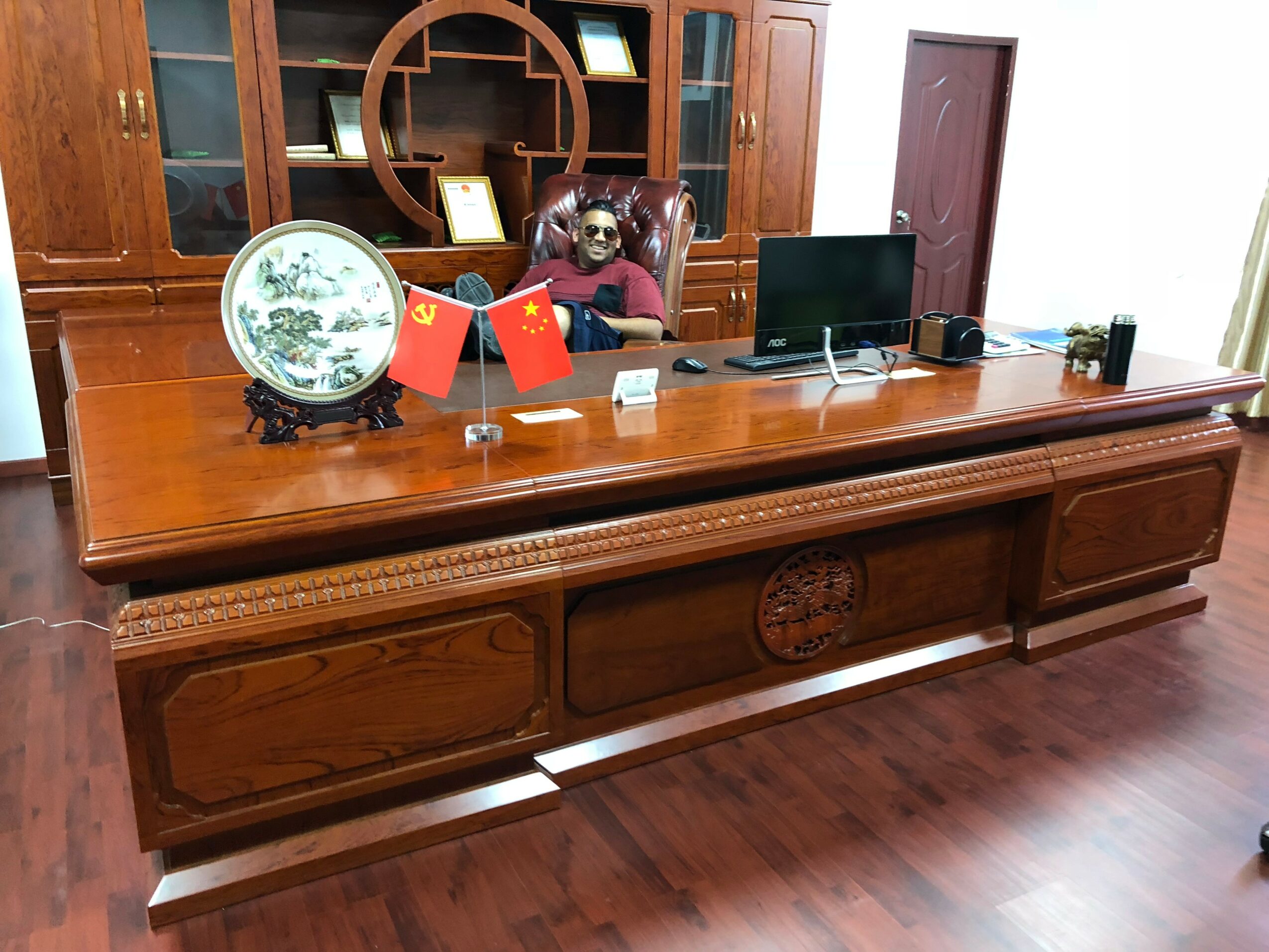Person sitting at a large wooden desk with a computer, flags, and decorative items, in a room with wooden furniture and flooring.