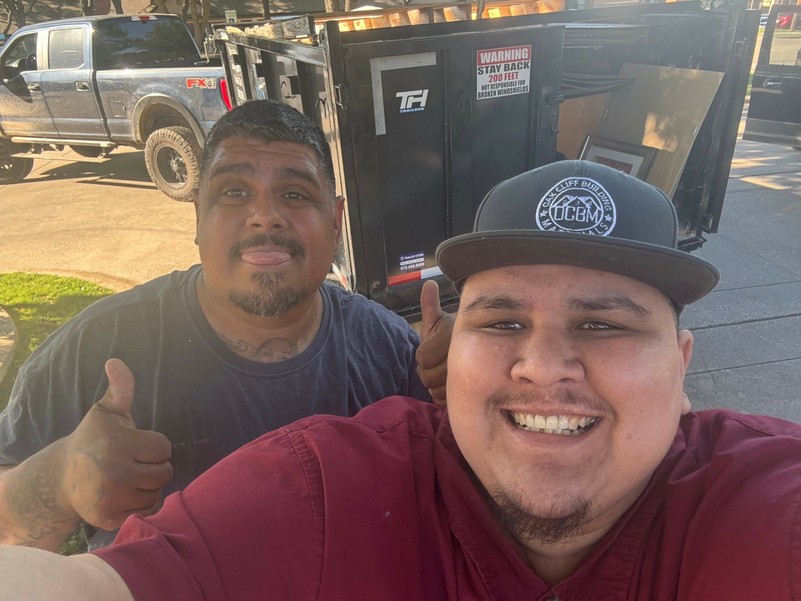 Two men smiling, one wearing a hat, outdoors with a vehicle in the background.