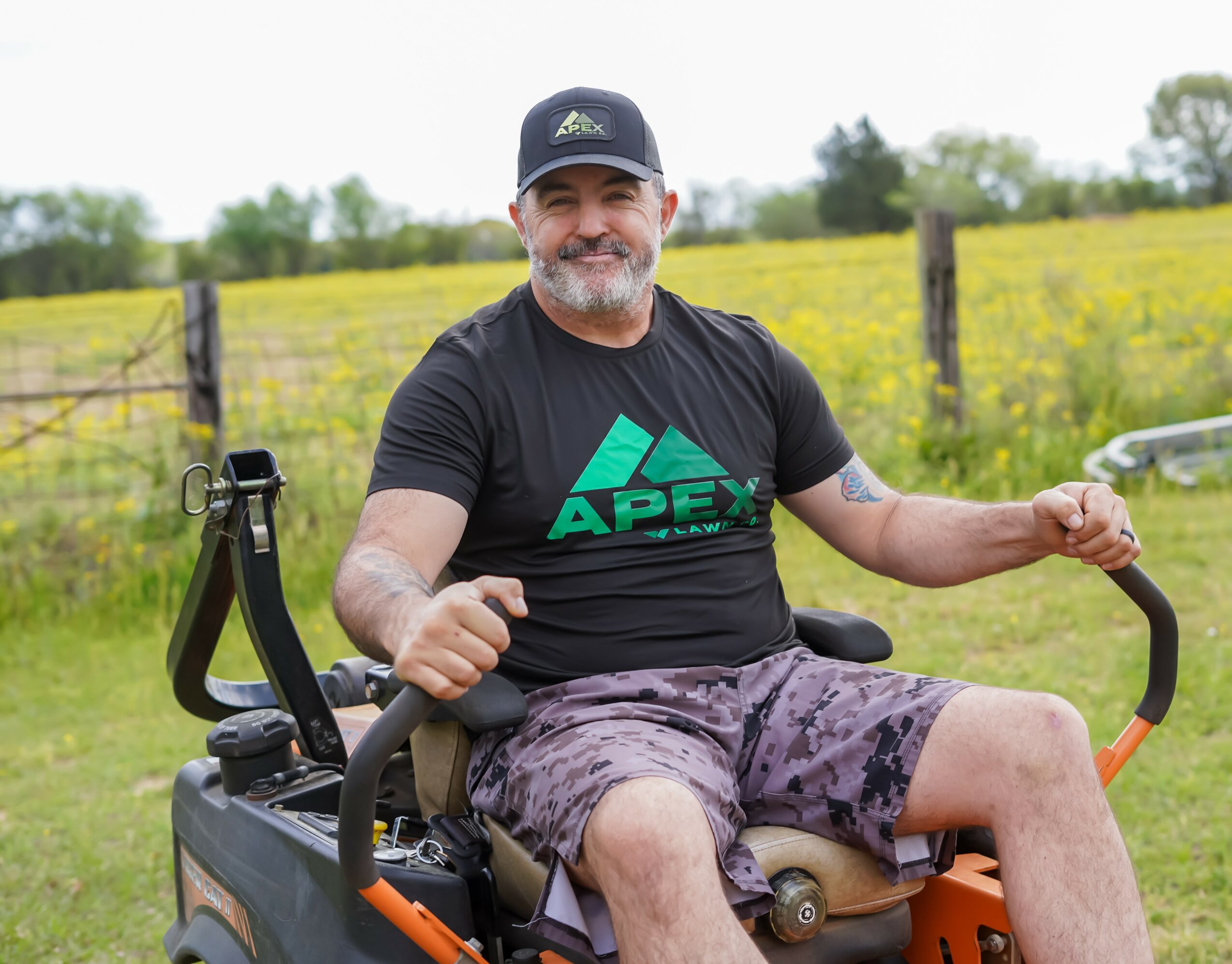 Smiling man with a beard and cap sitting outdoors on a mobility device, holding handlebars, with a field and fence in background.