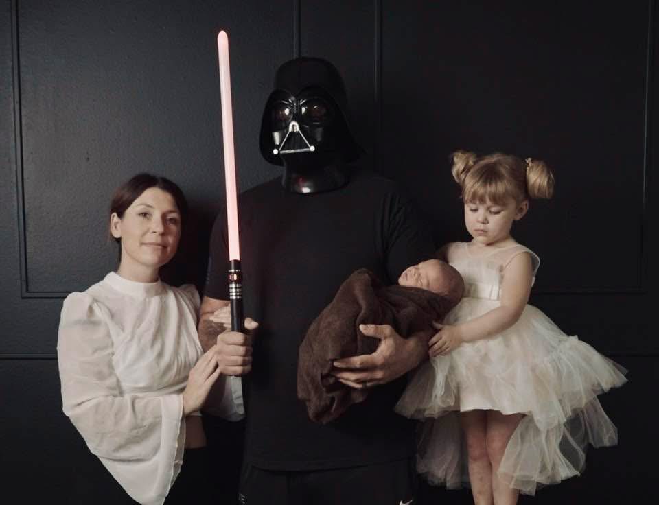 Person dressed as Darth Vader holding a child in a white dress, with a woman standing beside them, all against a dark background.
