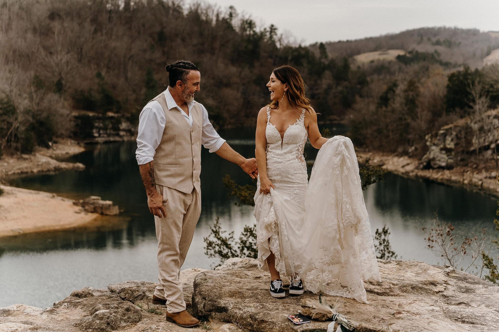 A couple holding hands outdoors near a river, with trees and hills in the background, dressed in wedding attire.