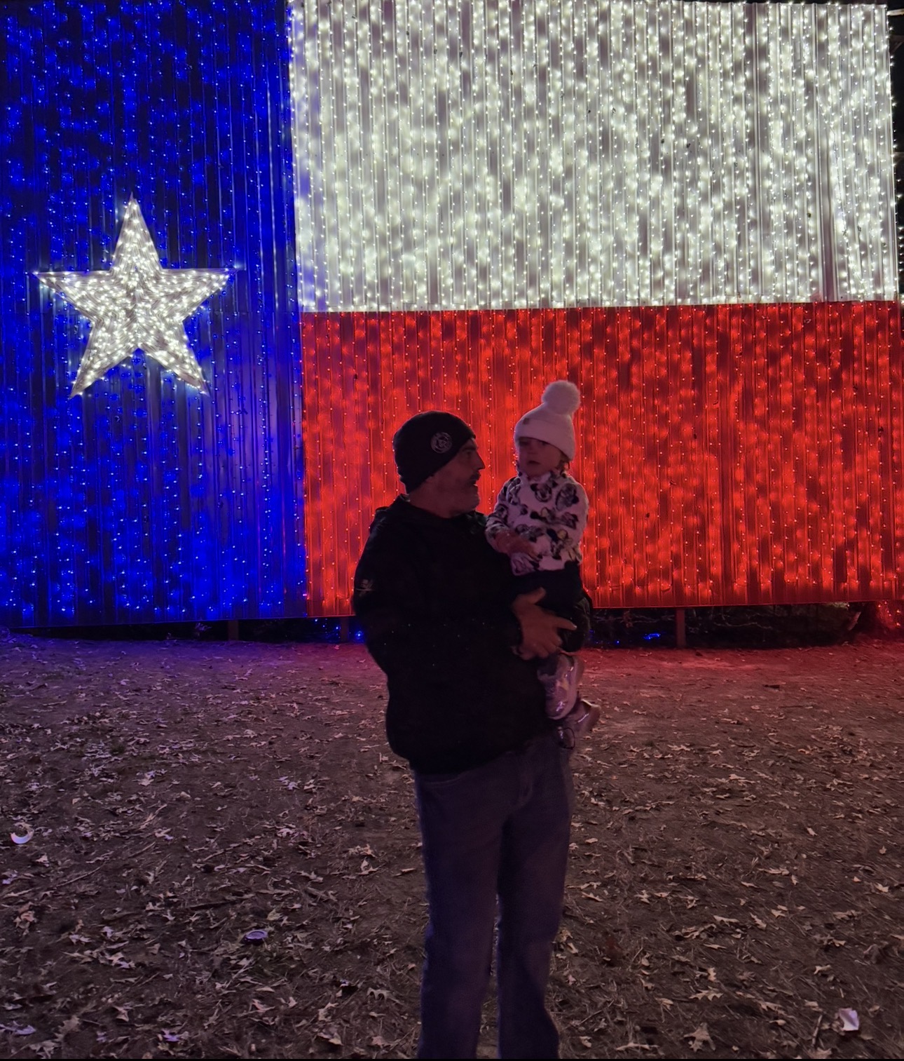 Person holding a child in front of a large illuminated flag with a star, red, white, and blue colors, at night.