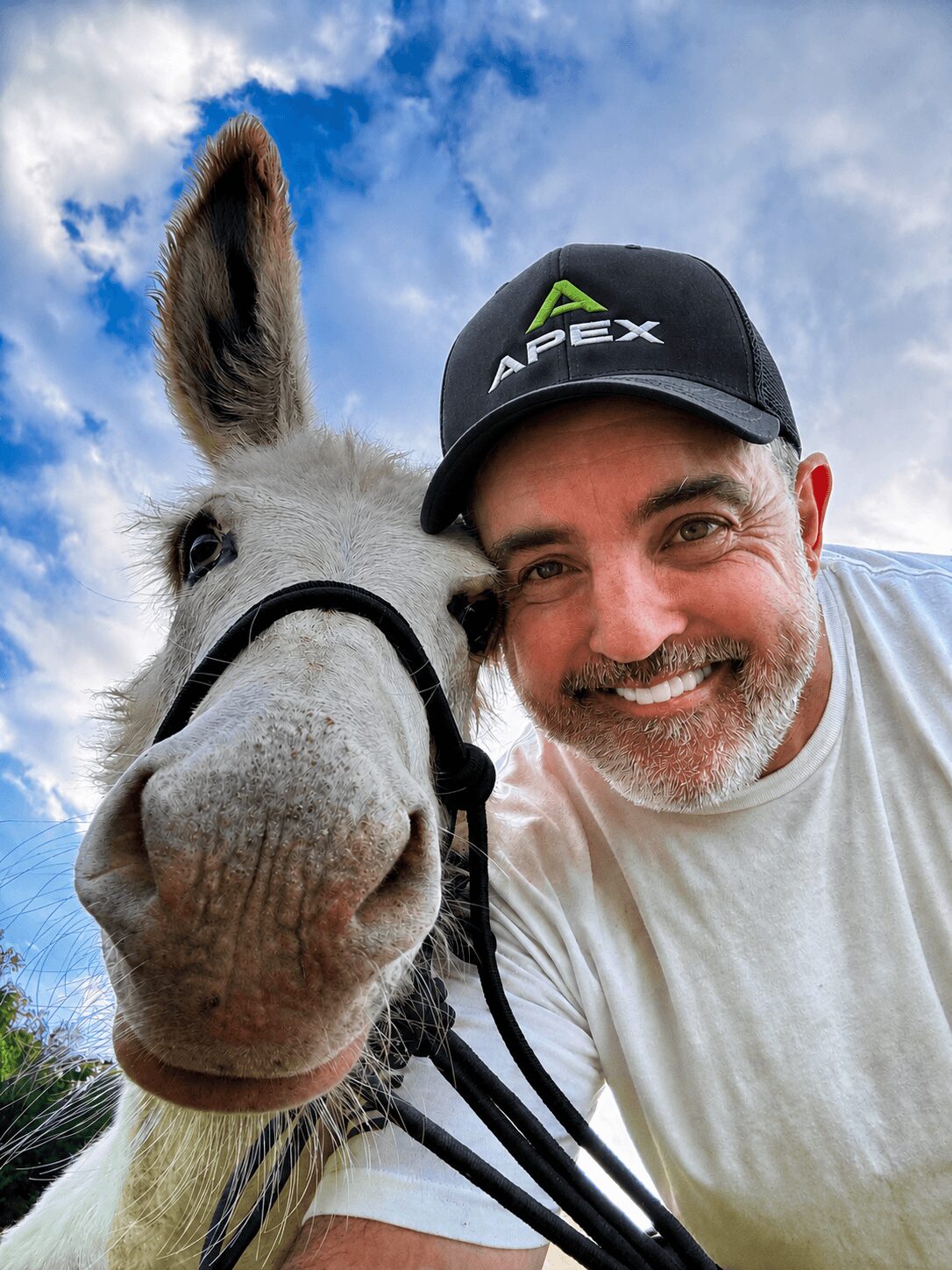 Man smiling next to a donkey outdoors with blue sky and clouds, wearing a black cap and white shirt.